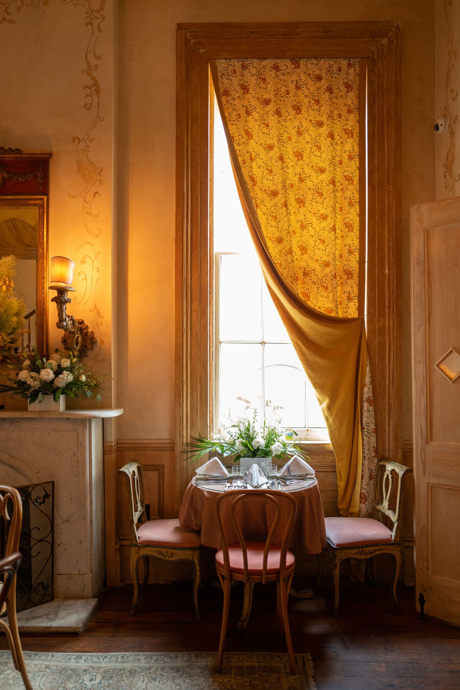 Elegant Victorian parlor room with ornate gold curtains, antique chairs, and natural light streaming through tall windows.