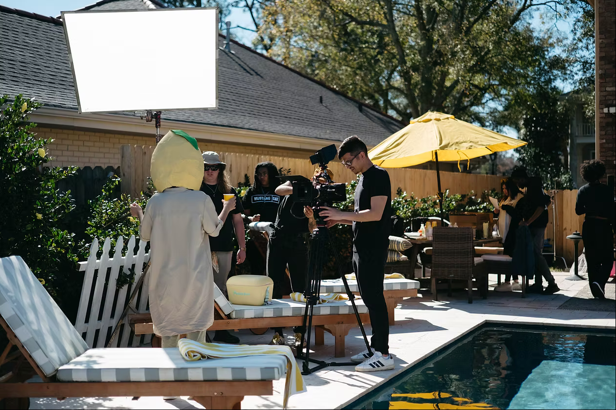 Film crew shooting a scene by a pool with camera equipment, yellow umbrella, and reflector board on a sunny day.