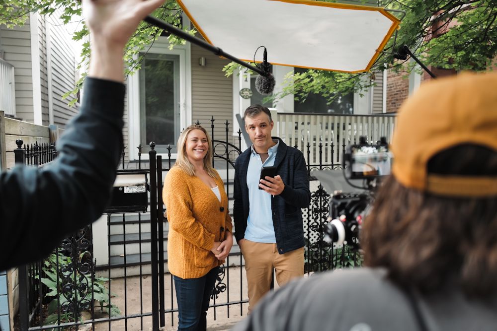 Film crew capturing a man and woman smiling outside a residential home with a black iron fence.