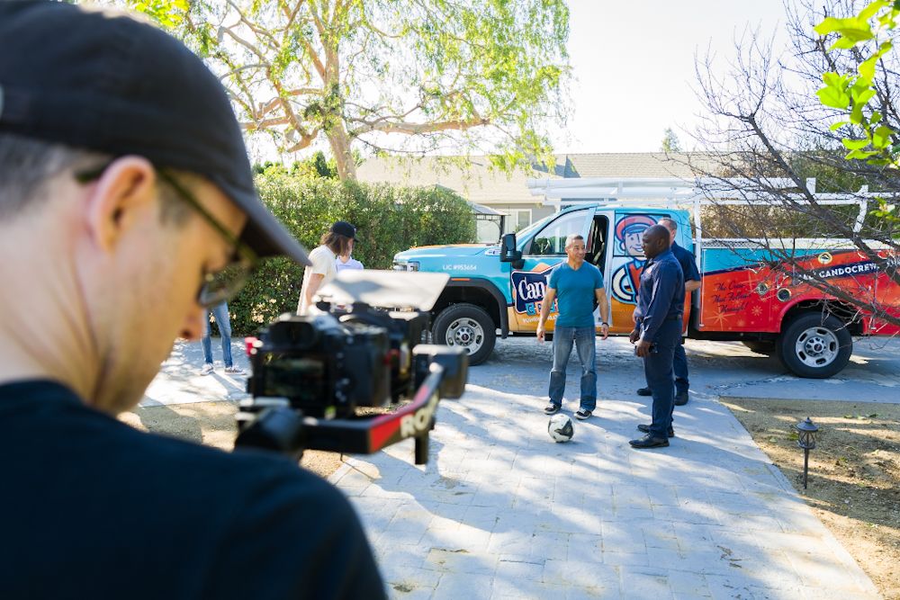 Filmmaker with camera filming a outdoor scene with people standing near colorful wrapped trucks on a sunny day.