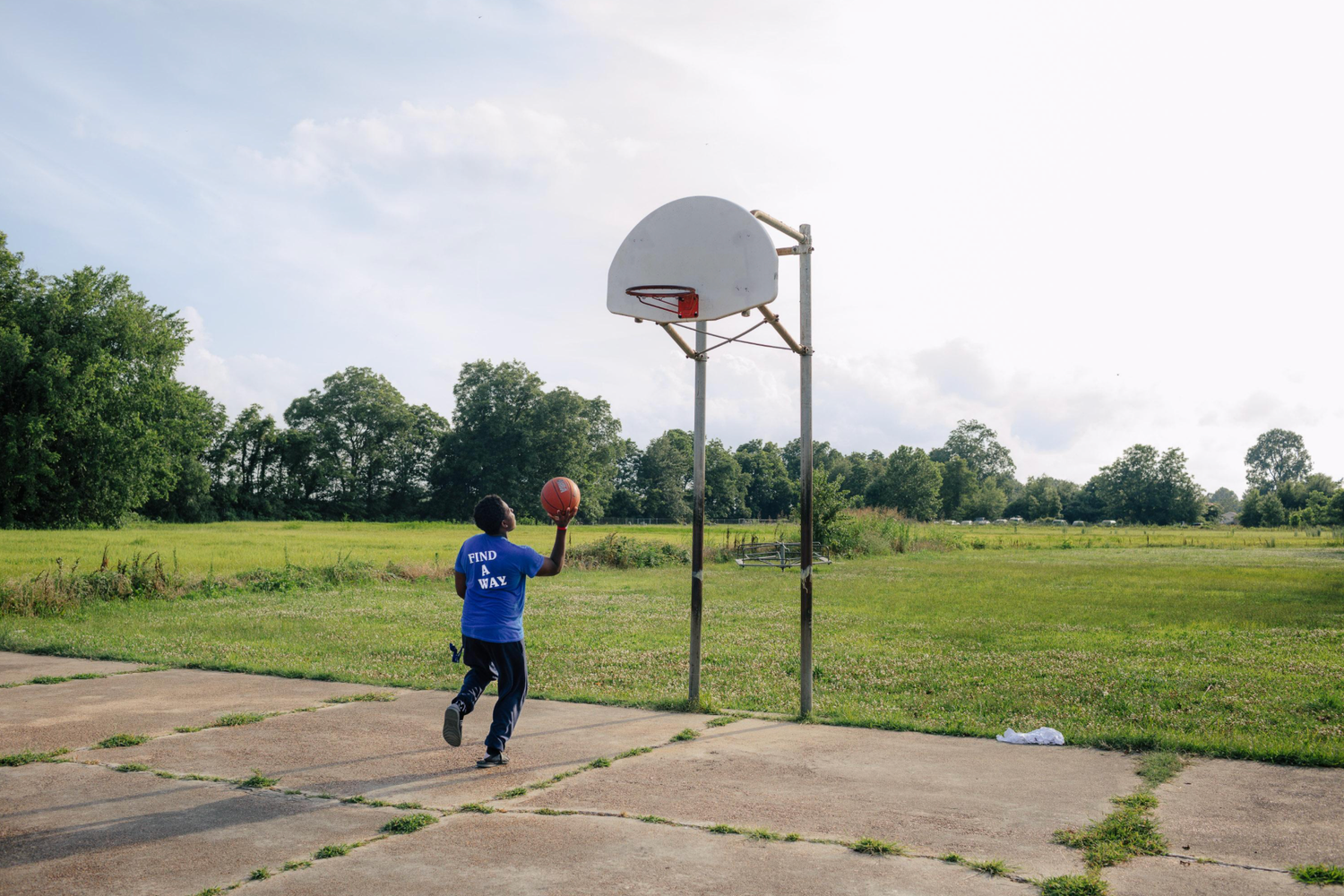 A sequence of basketball players practicing jump shots on an outdoor court surrounded by green fields and trees.