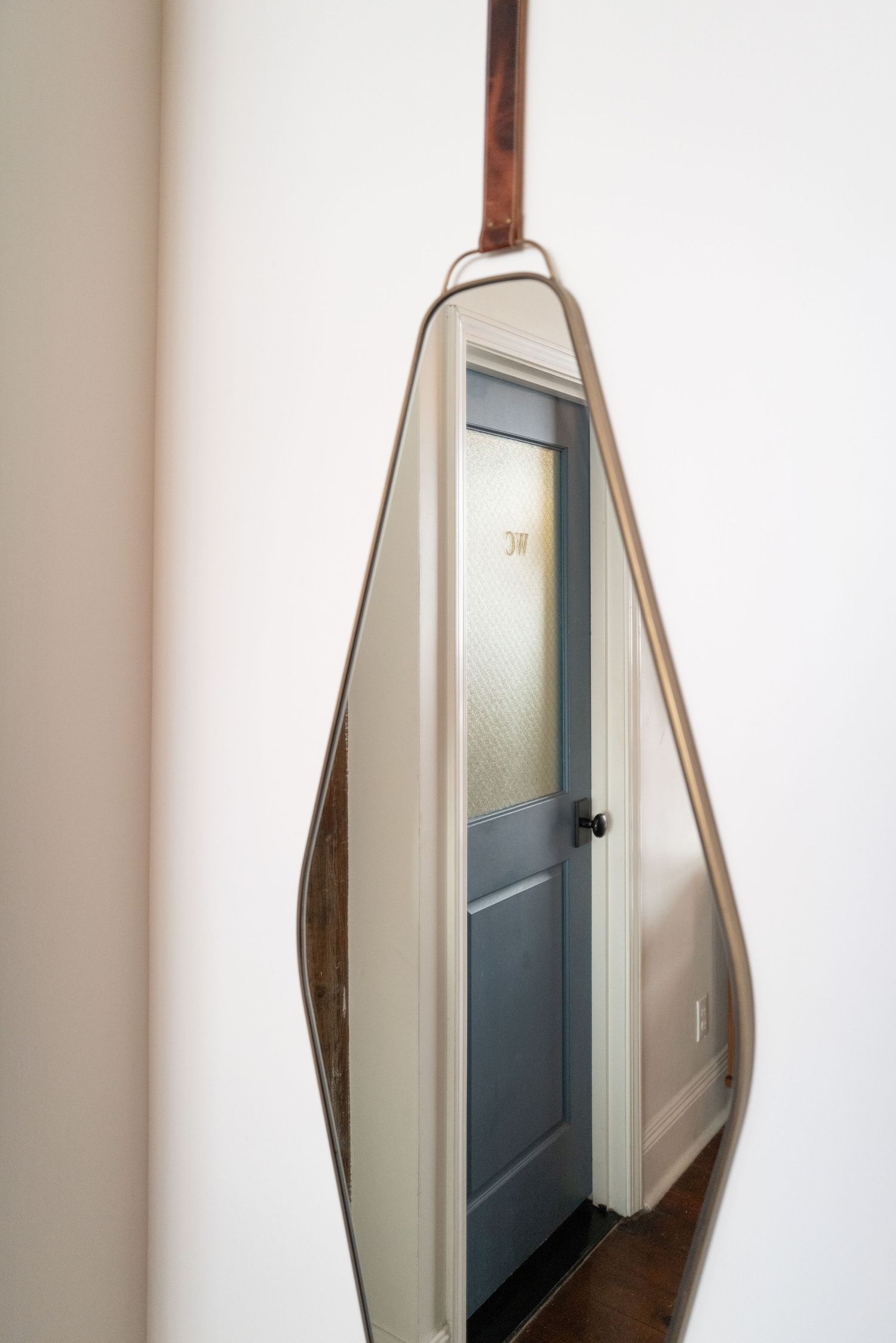 Teardrop-shaped mirror with leather strap hanging on white wall near blue door.