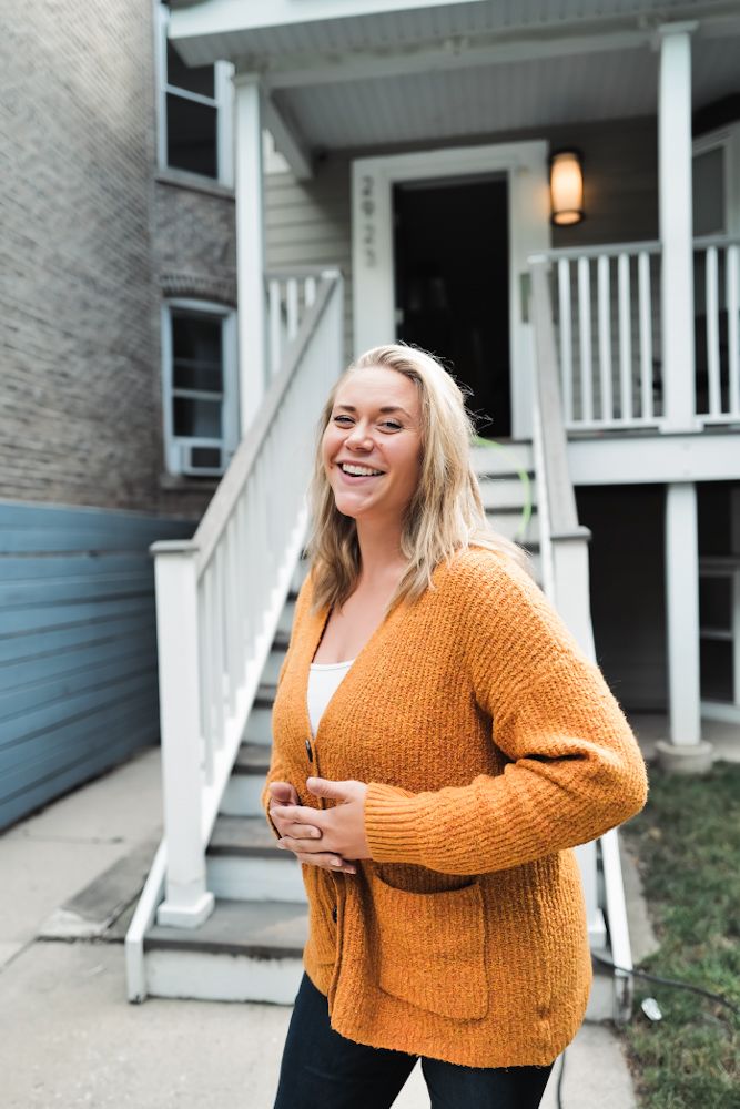 Smiling blonde woman in orange cardigan standing outside a brick apartment building with white staircase.