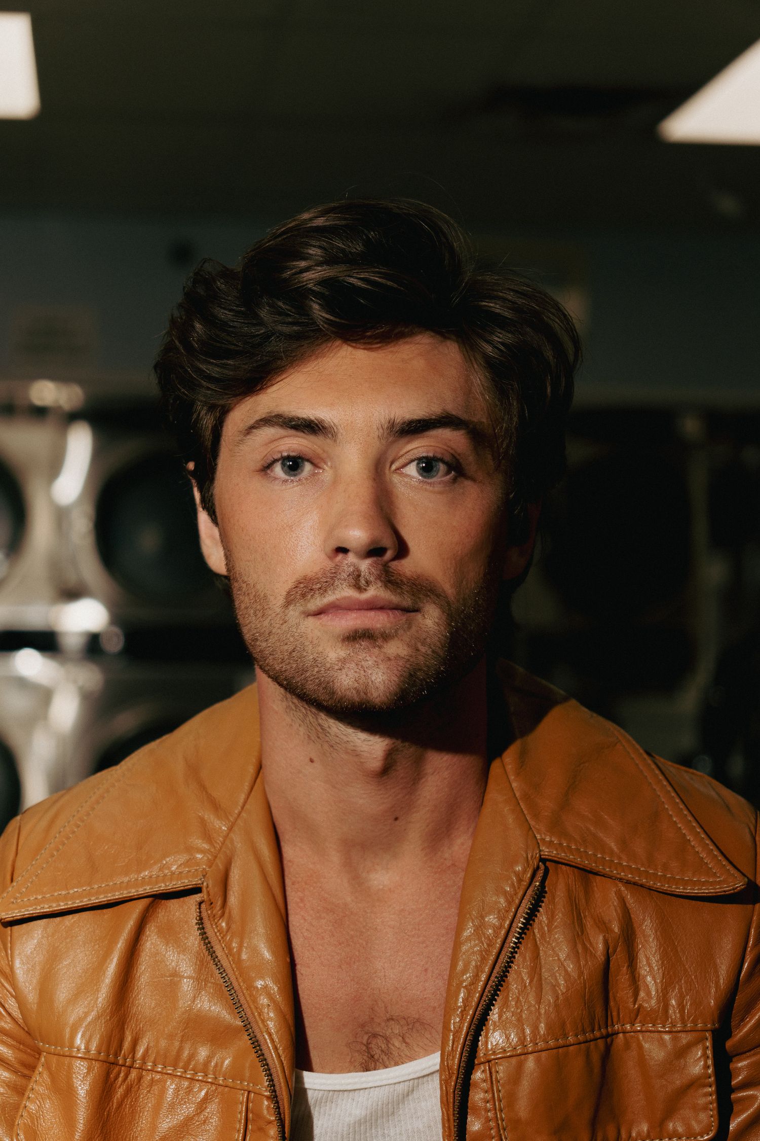 Young man with dark hair in a brown leather jacket poses seriously in a dimly lit laundromat.