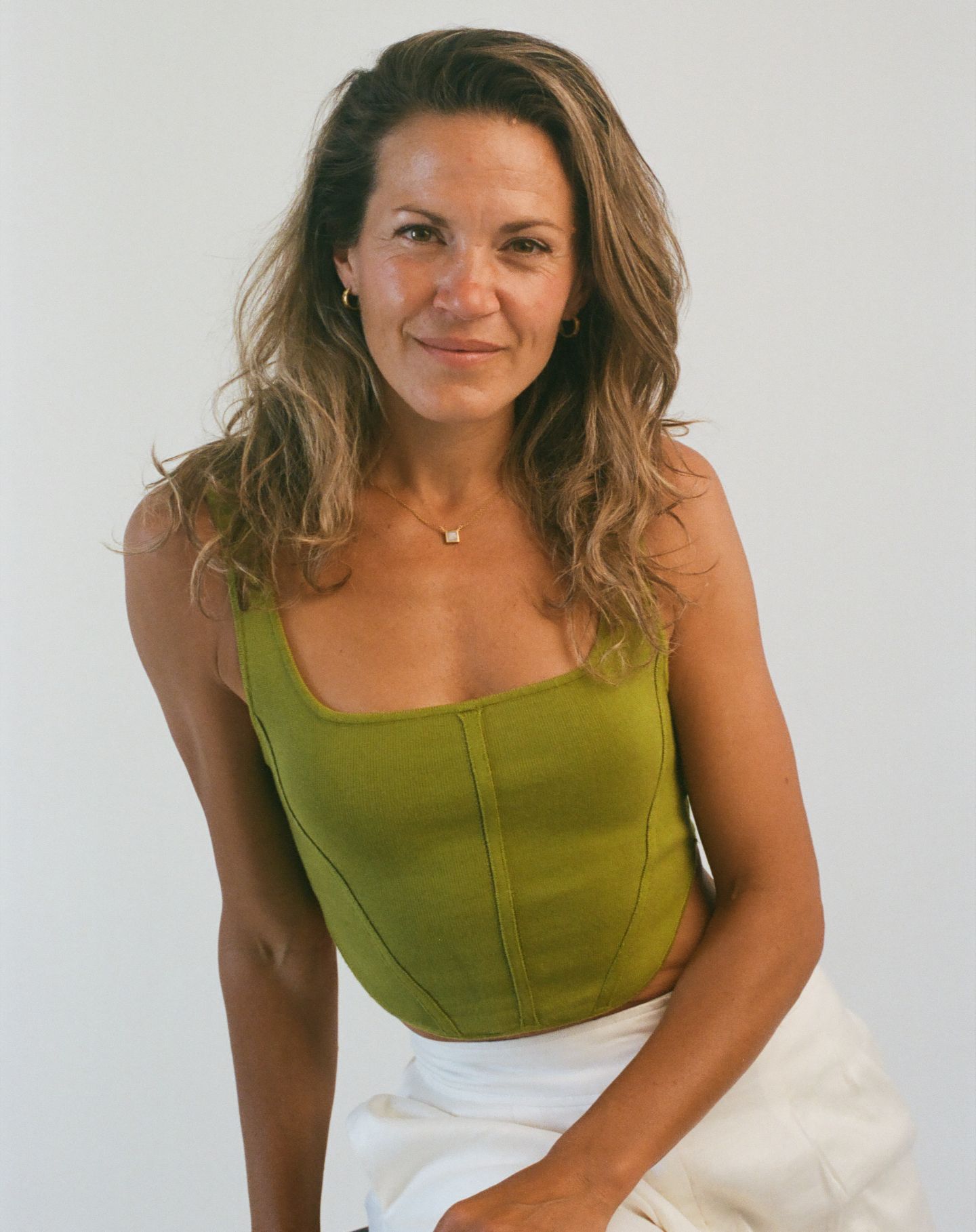 Person in olive green tank top against white background smiling with wavy brown highlighted hair.