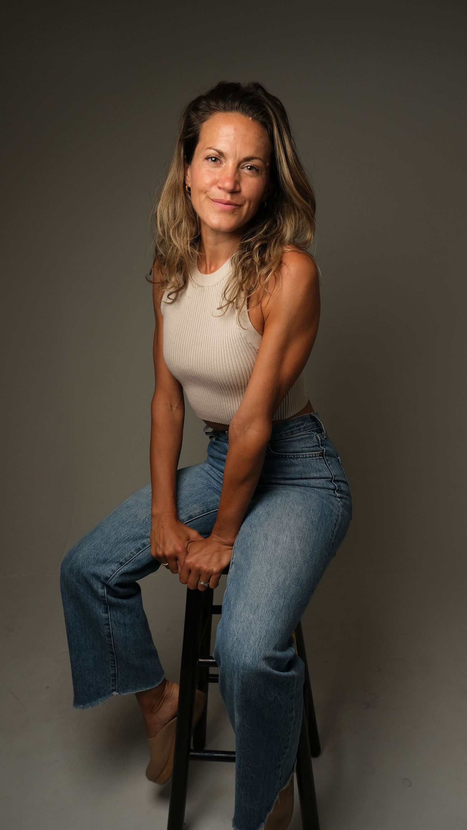 A woman in a gray tank top and jeans poses on a stool against a dark backdrop in a studio photoshoot series.