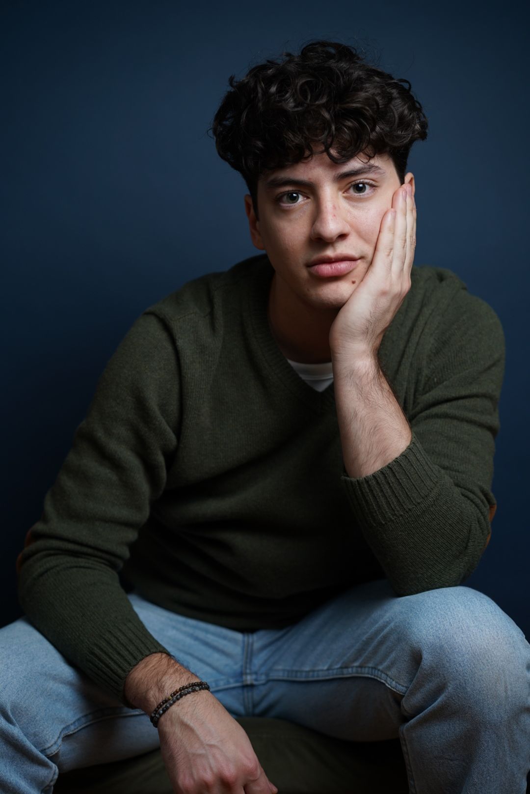 A young person in a green sweater and jeans poses casually against a dark background in a photo series.
