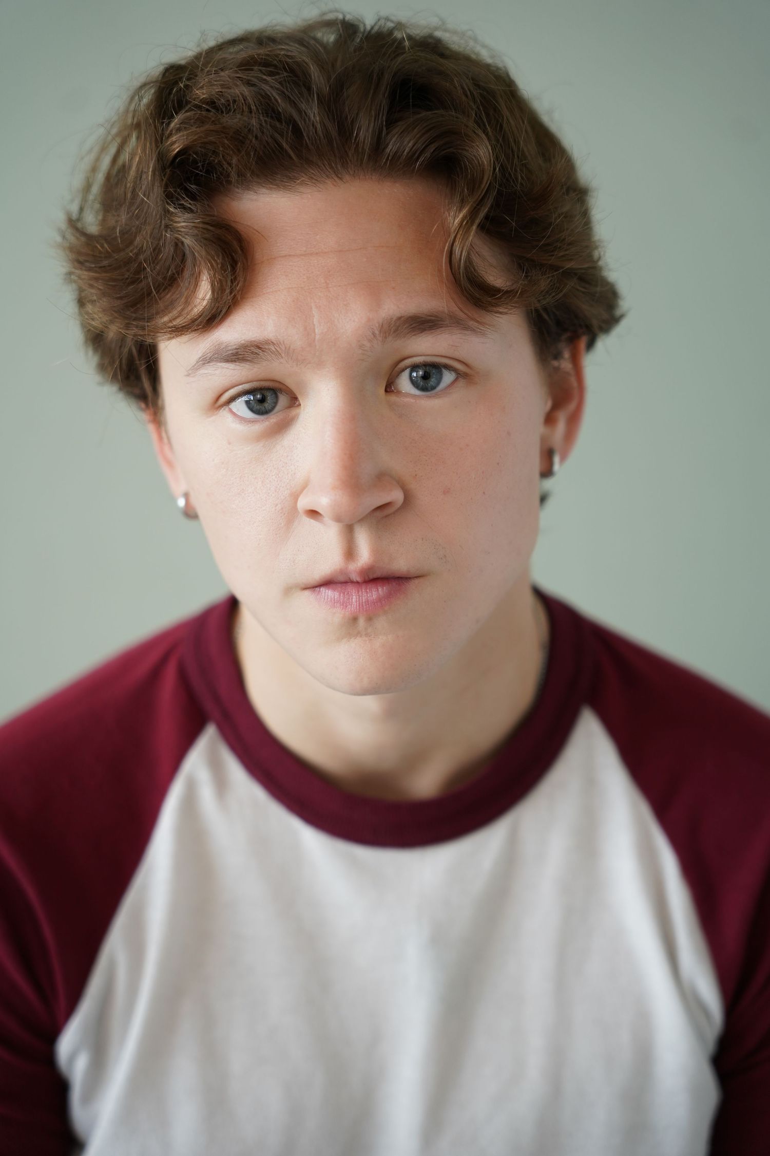 Young person with curly brown hair and blue eyes wearing a maroon and white raglan t-shirt against a light background.