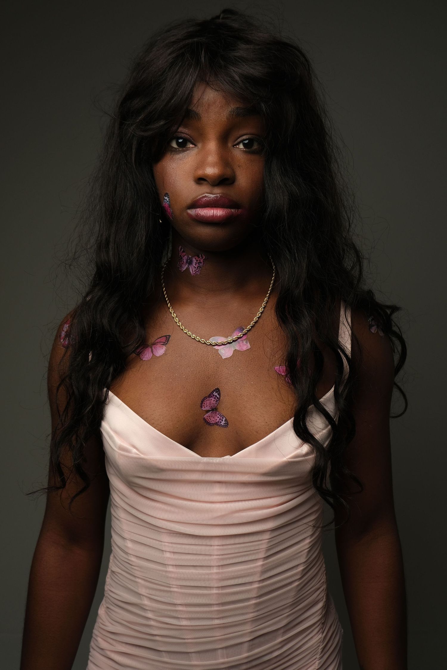 A portrait studio shot of someone in a pale pink ruched dress with long dark wavy hair and a butterfly decoration.