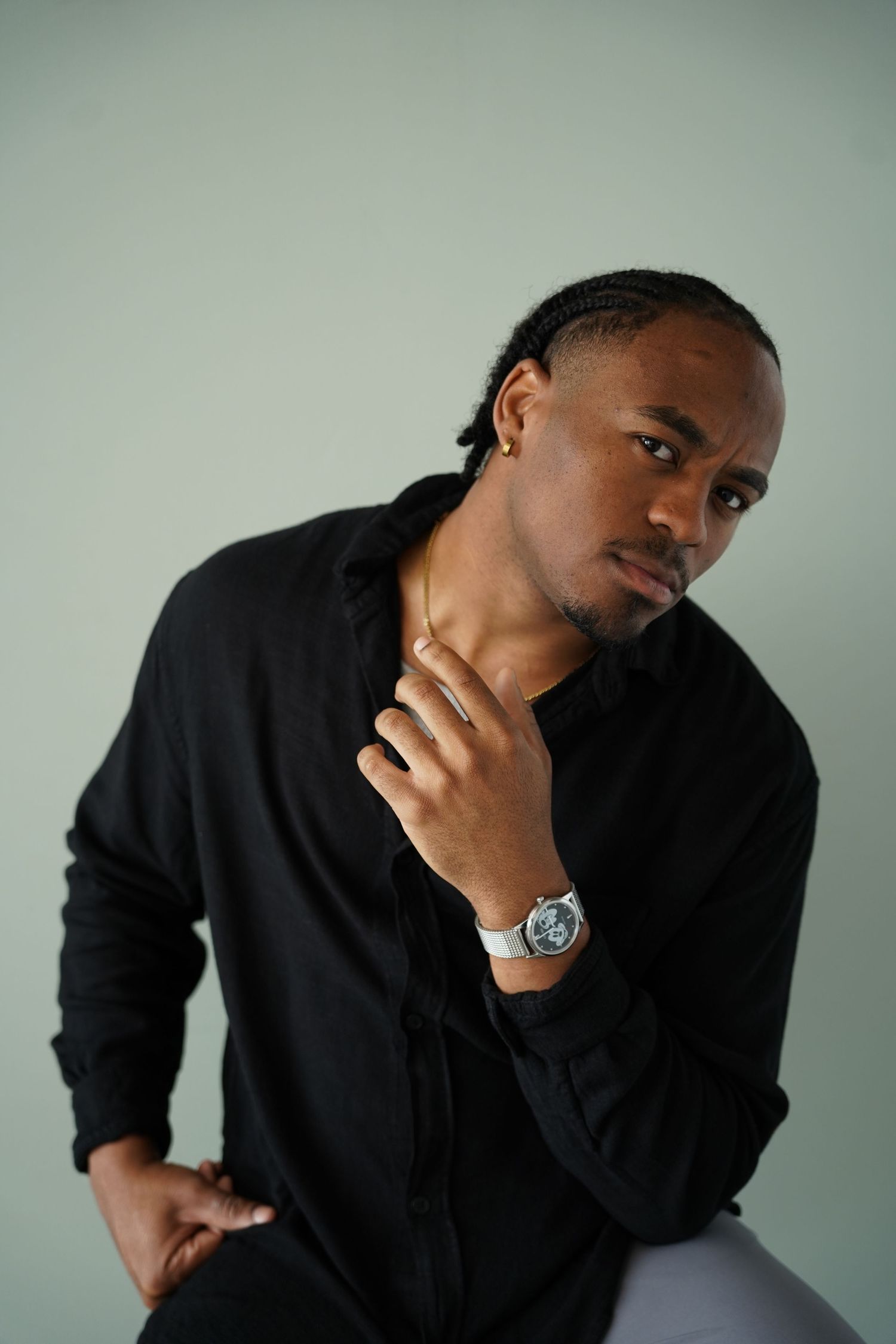 A young Black man in a black shirt and silver watch sits confidently against a light green background.
