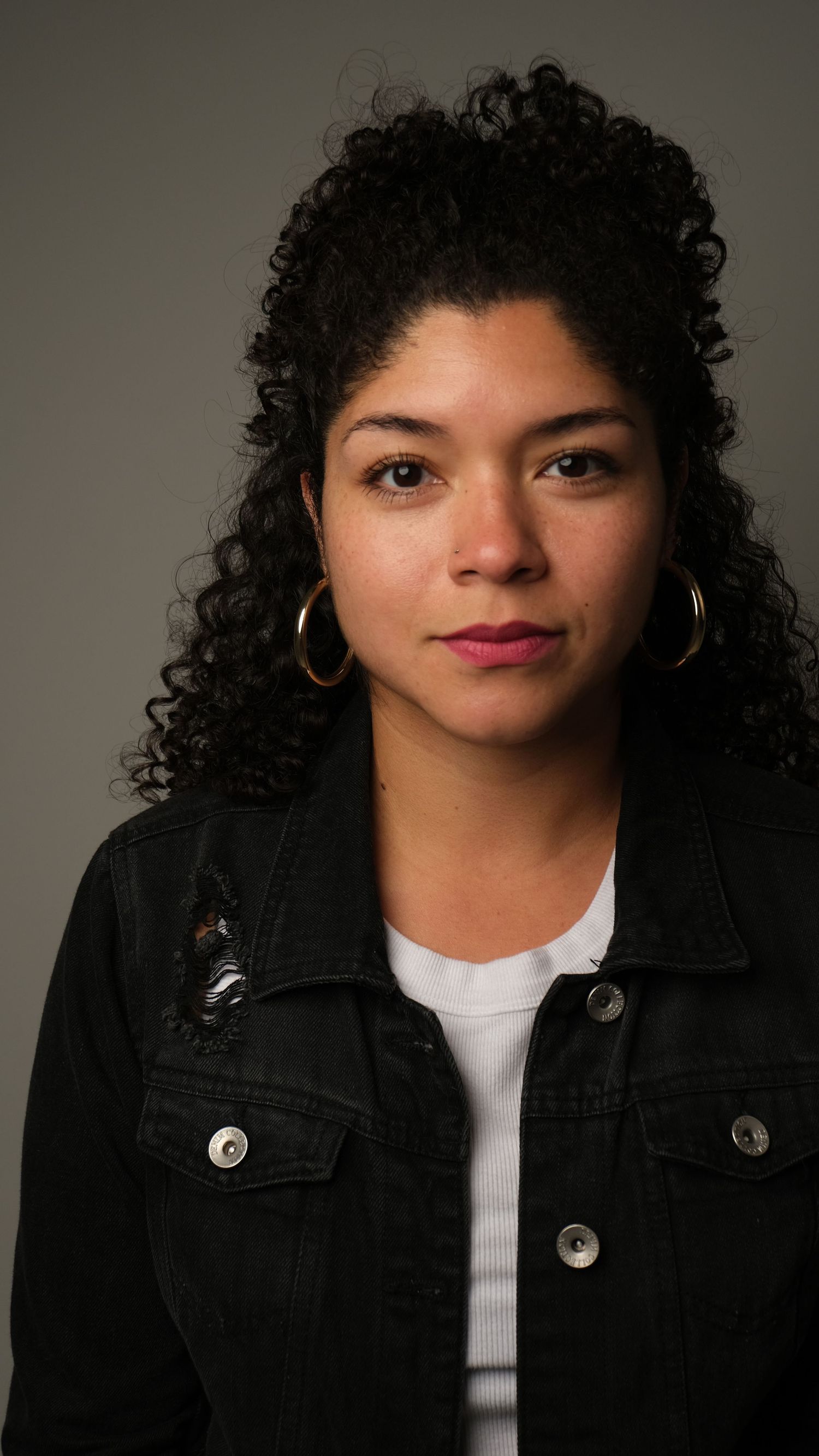 Portrait of a woman with curly dark hair wearing a black denim jacket and silver hoop earrings against a neutral background.