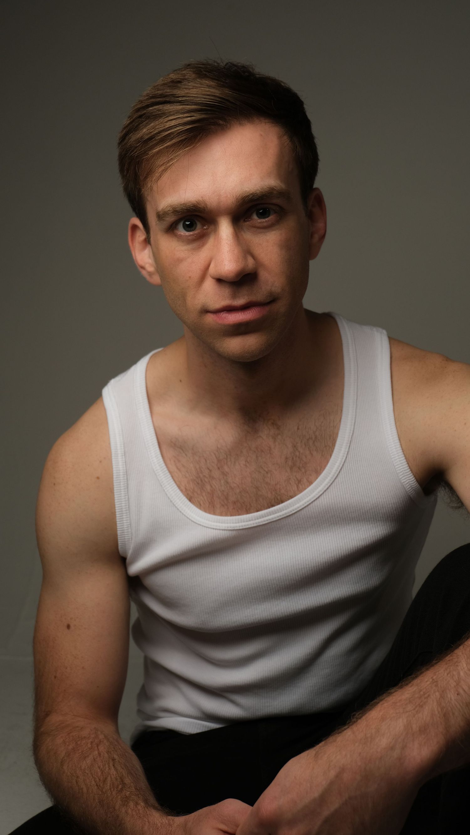 Studio portrait of a young person in a white tank top against a dark background.