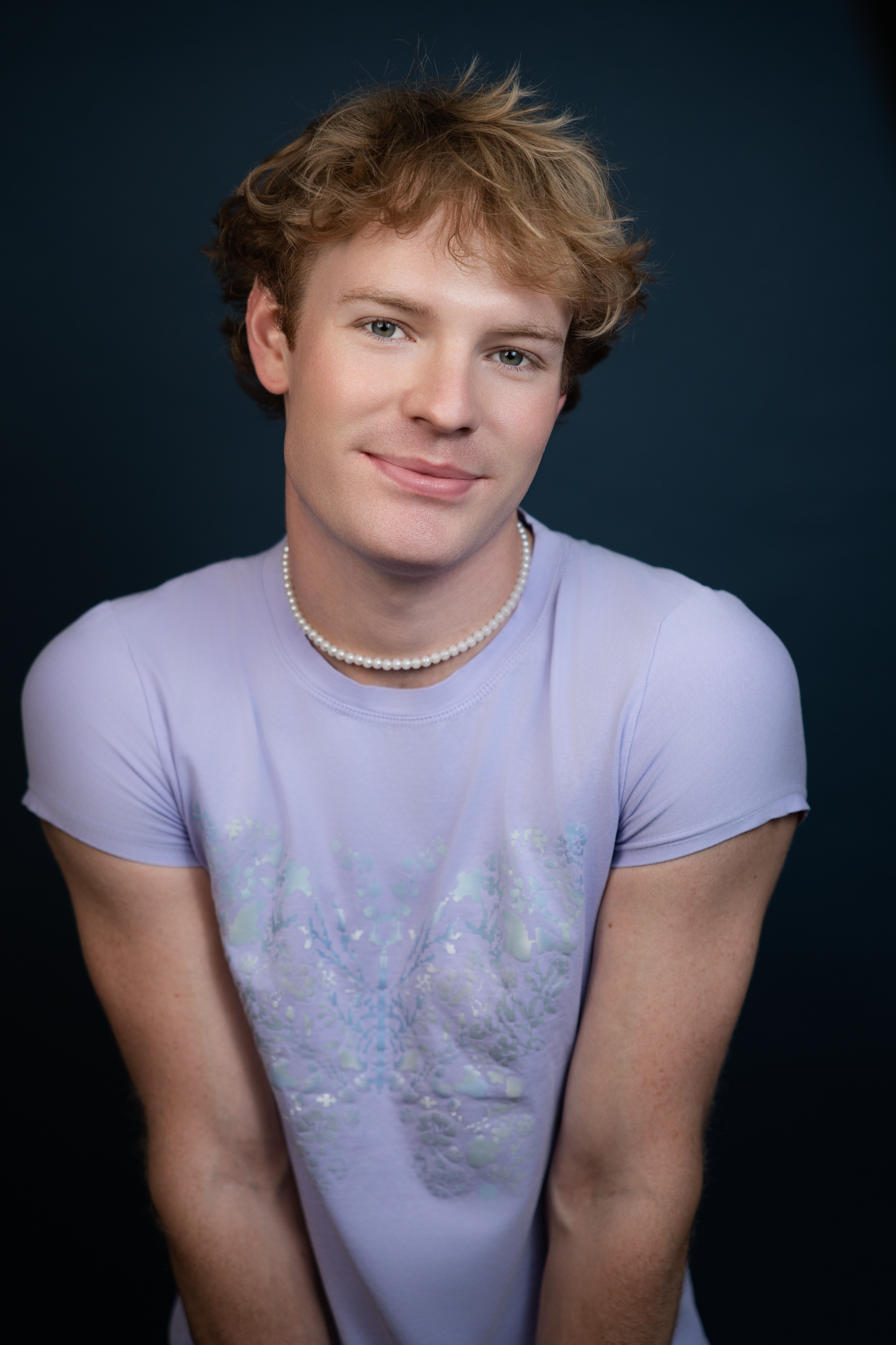 Young man with blonde hair wearing a lavender t-shirt and pearl necklace, smiling against dark background.