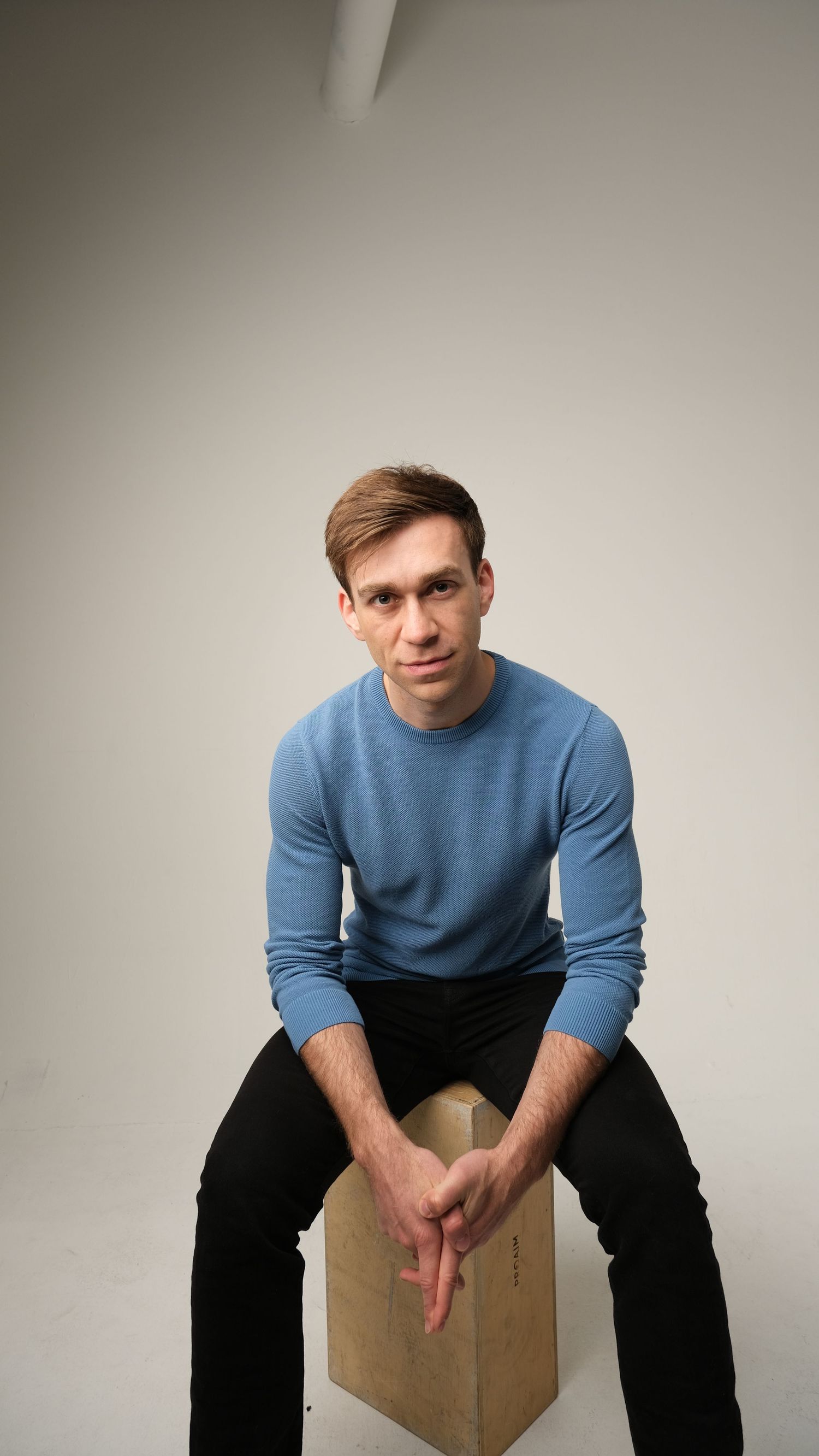 Person in light blue sweater and dark pants sitting on wooden box against white background.