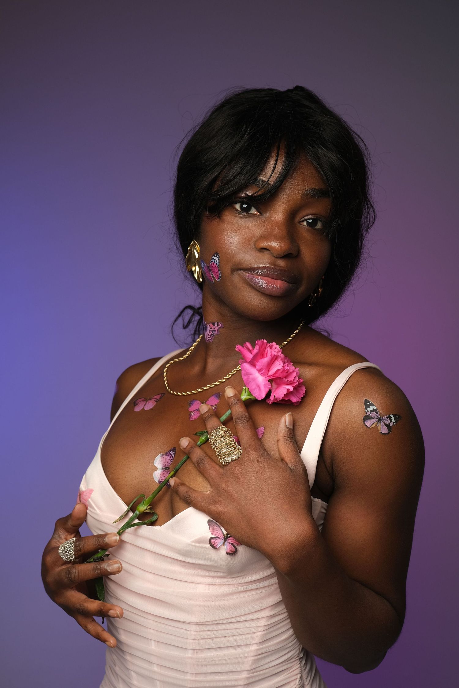 Woman in pink dress holding pink flower against purple background, wearing gold jewelry and butterfly tattoo.