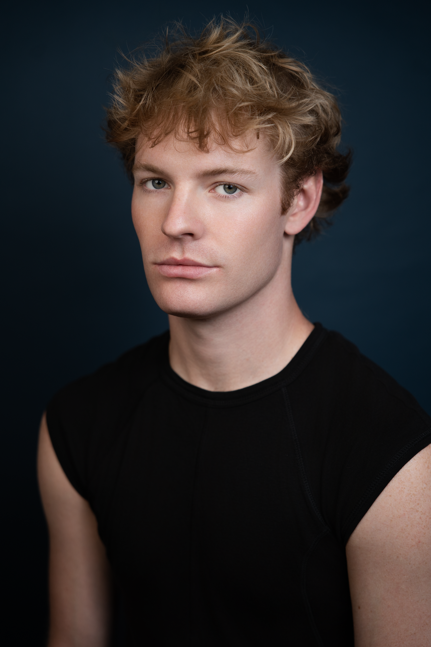 Young man with curly auburn hair wearing a black sleeveless shirt poses against a dark blue studio background.