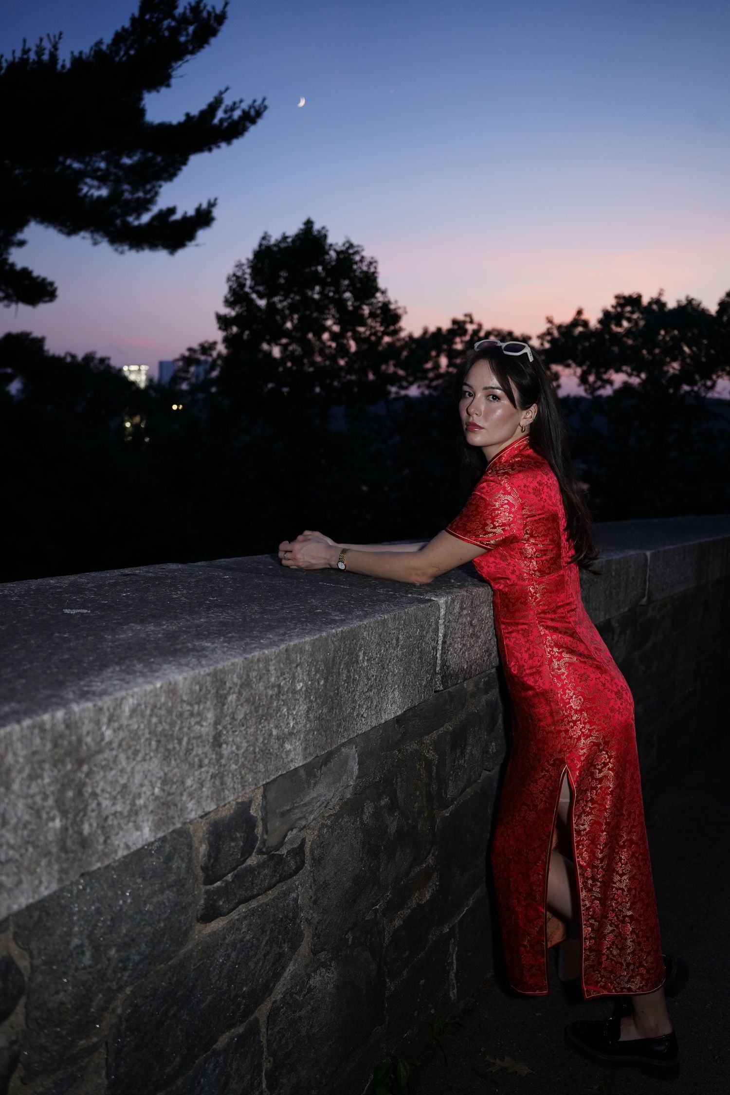 Woman in red cheongsam dress leaning on stone wall at dusk with crescent moon and colorful sky.