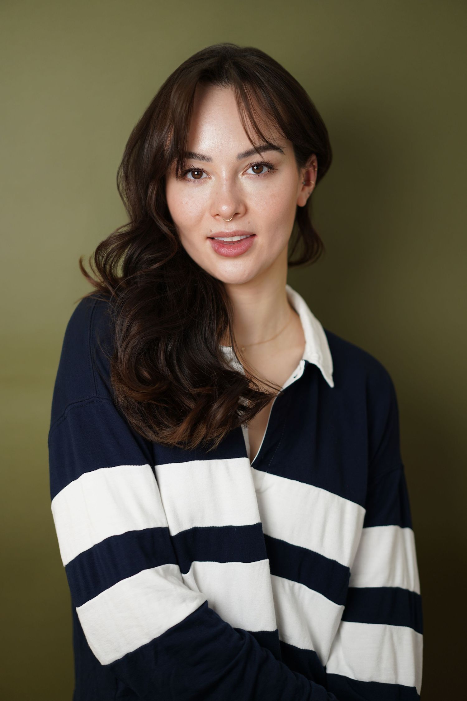 Young woman with dark wavy hair wearing a navy striped shirt, posing against an olive green background.