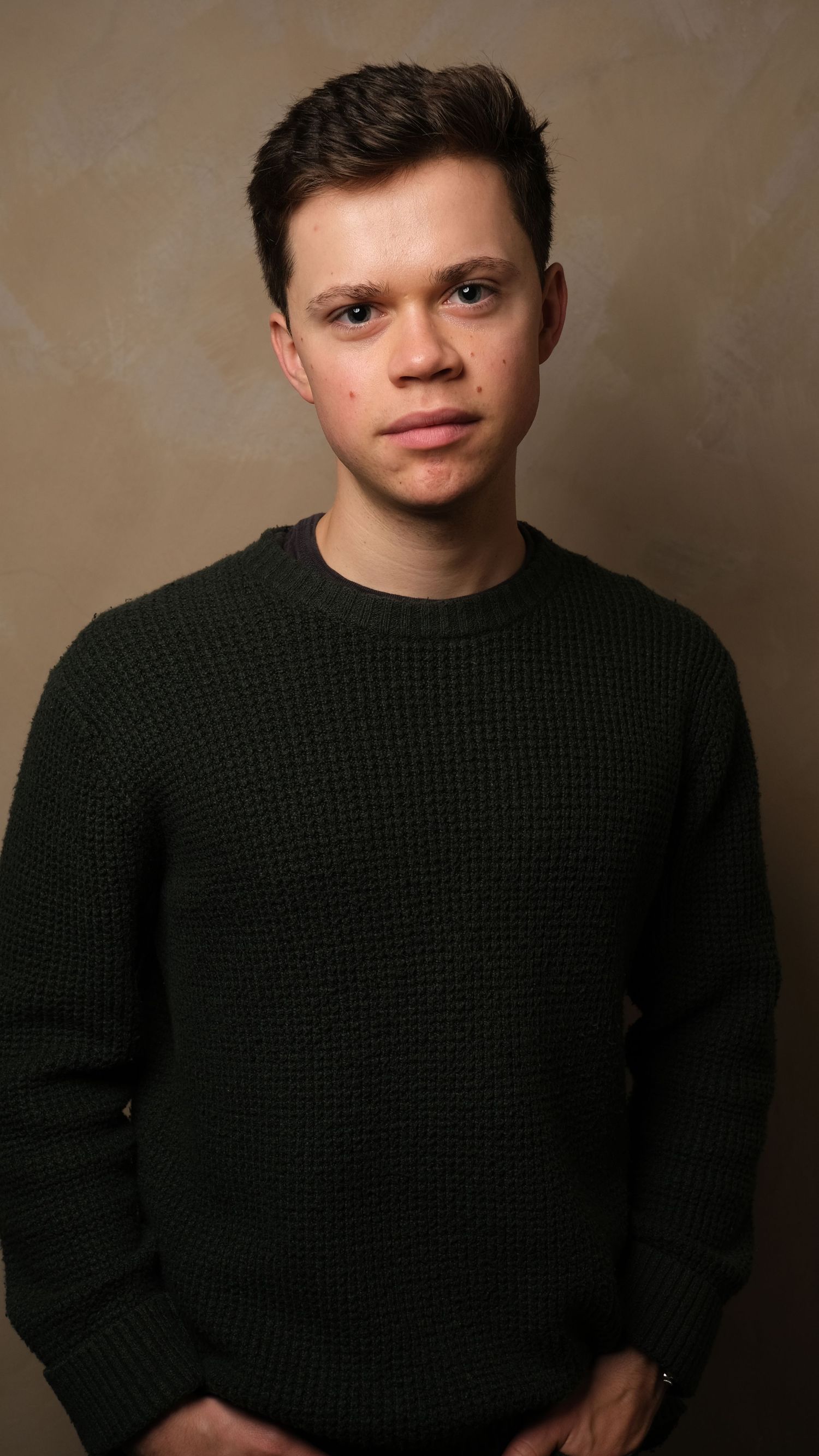A young person in a dark knit sweater poses against a neutral studio background in a portrait-style photo.