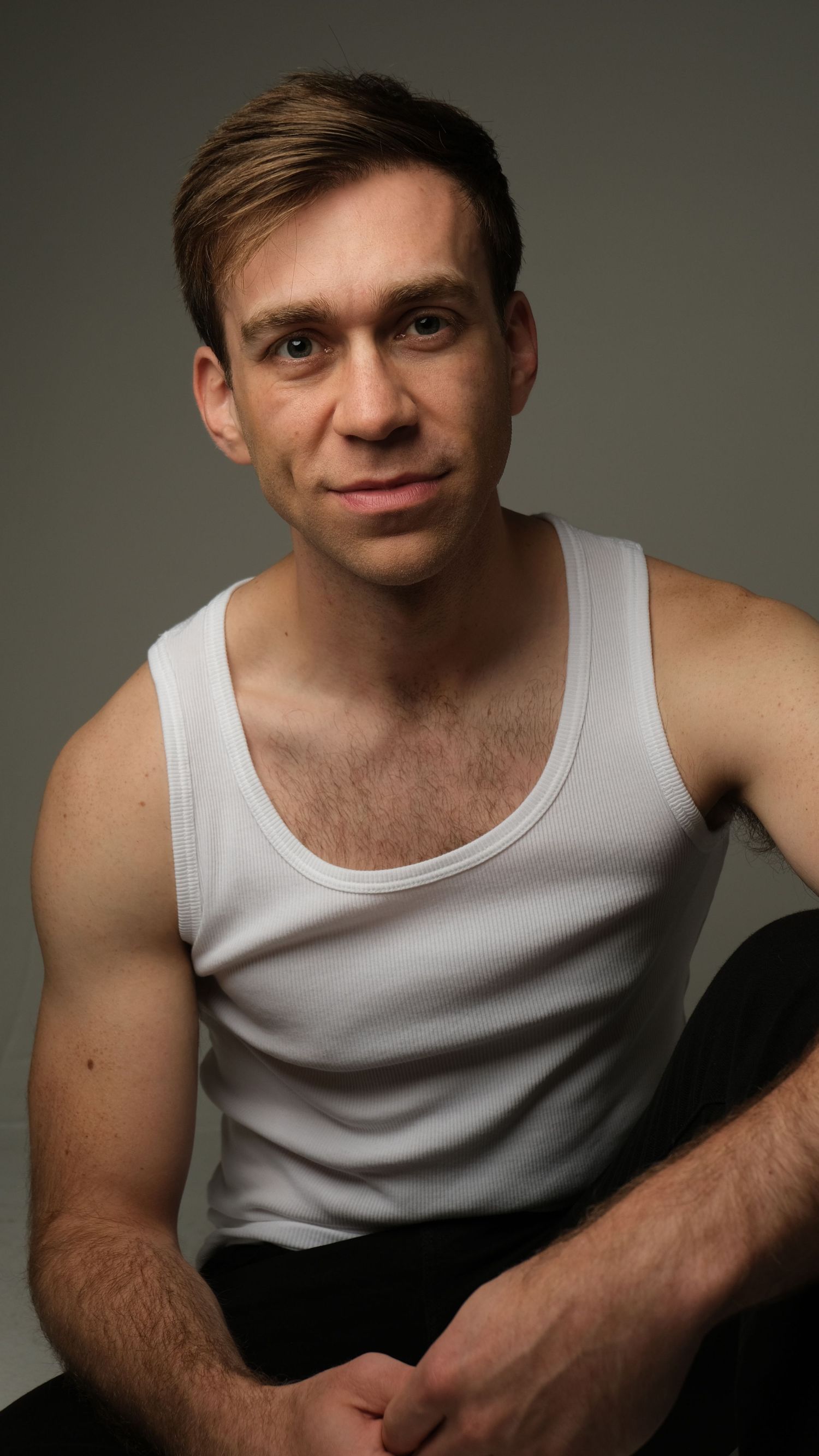 Portrait of a person wearing a white tank top sitting against a dark background.