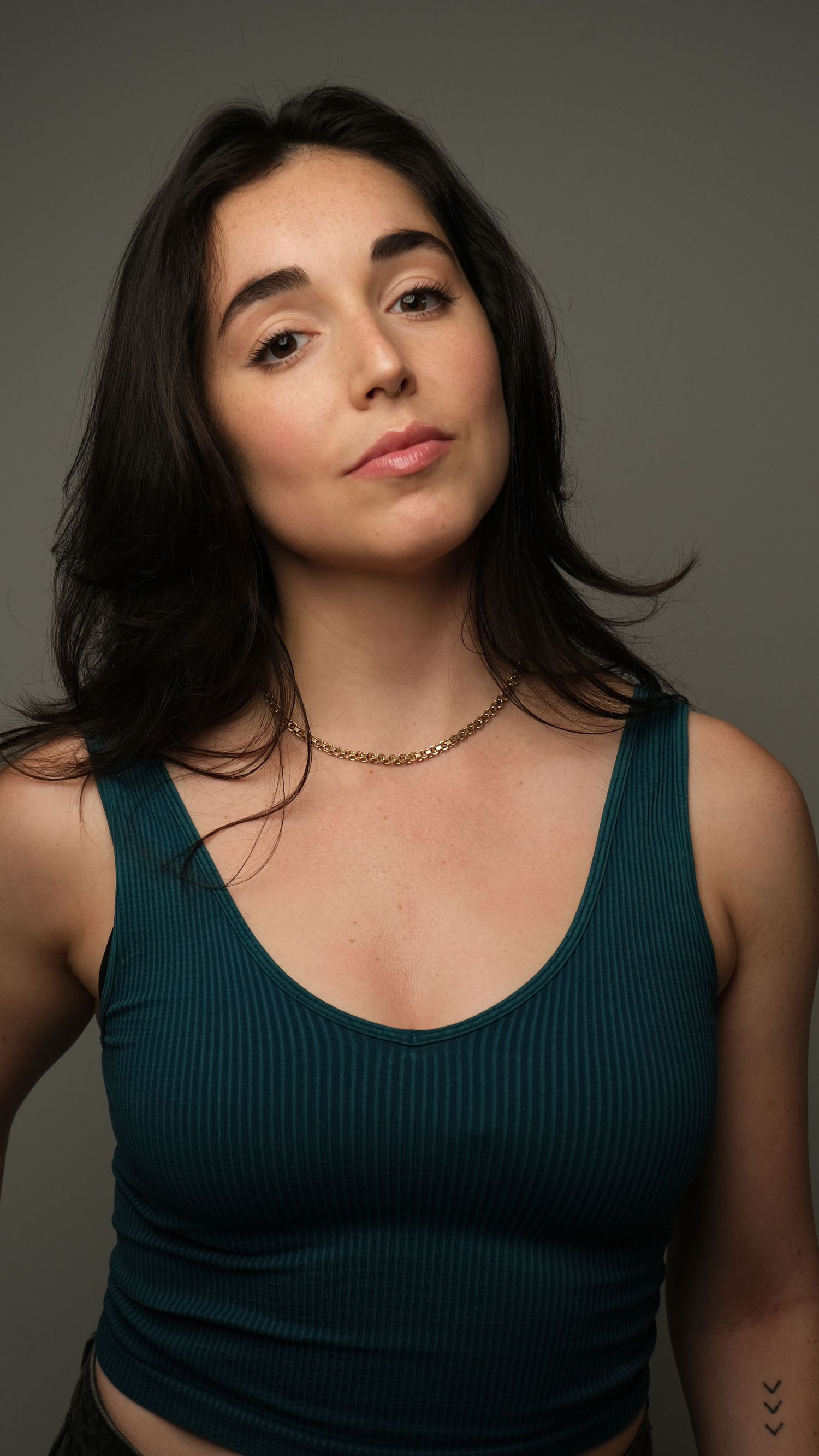 Studio portrait of a person wearing a dark teal tank top and gold necklace against a neutral backdrop.