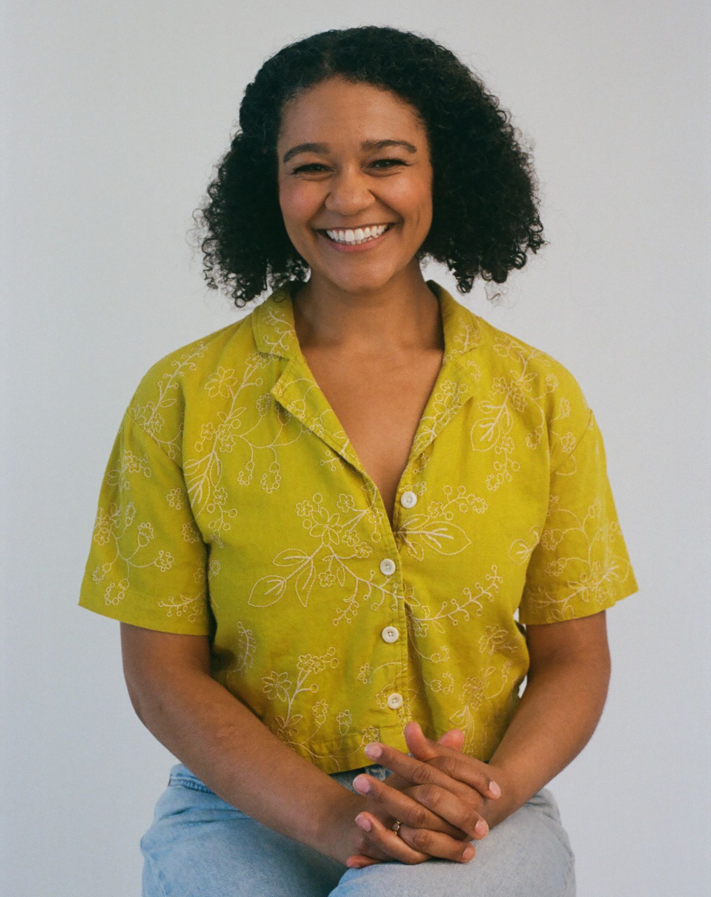 Woman in yellow floral button-up shirt and light jeans smiling and posing in four casual portraits against white wall.
