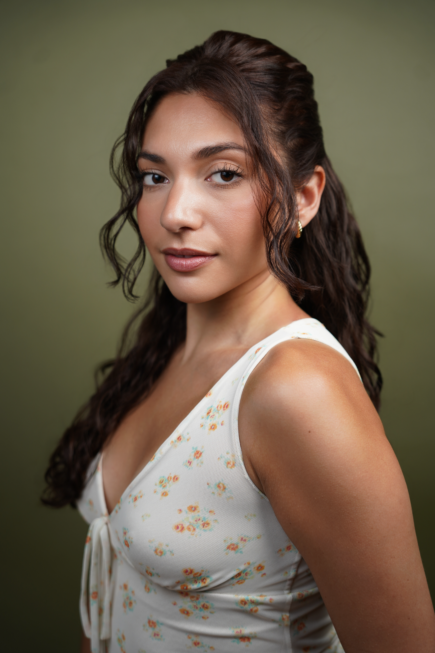 Young woman with dark wavy hair wearing a white floral sundress, posing against an olive green background.