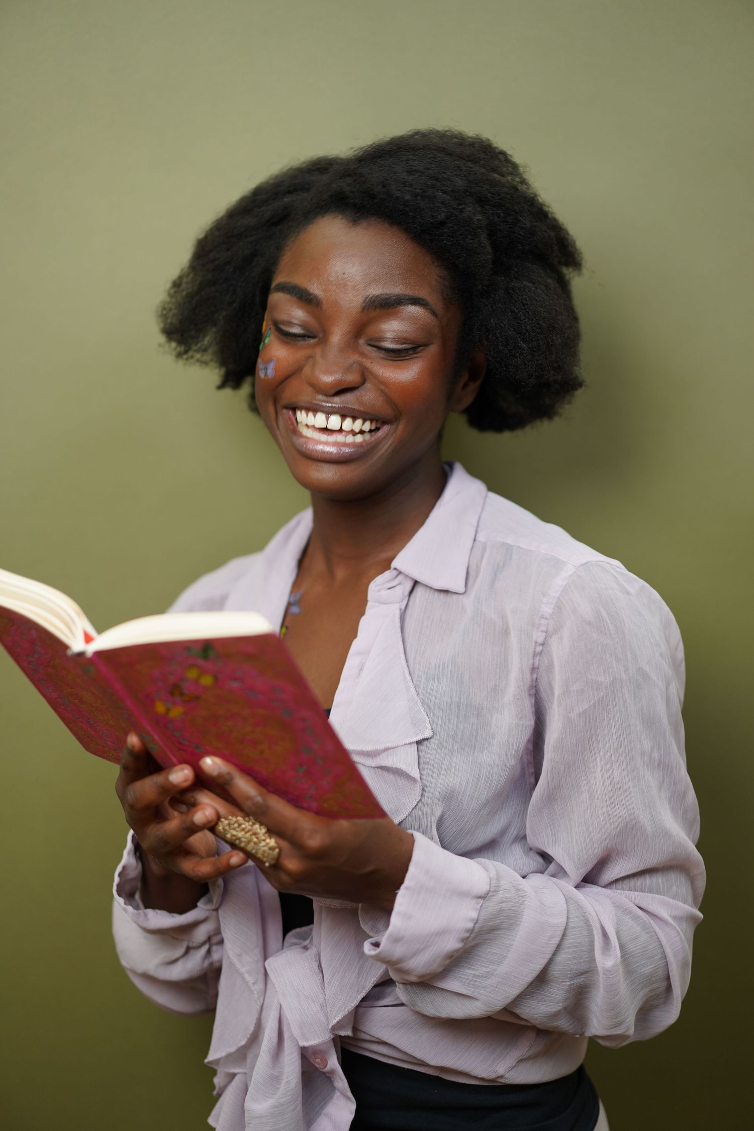 Smiling young Black woman with afro hair reading a red book, wearing a lavender blouse against an olive background.