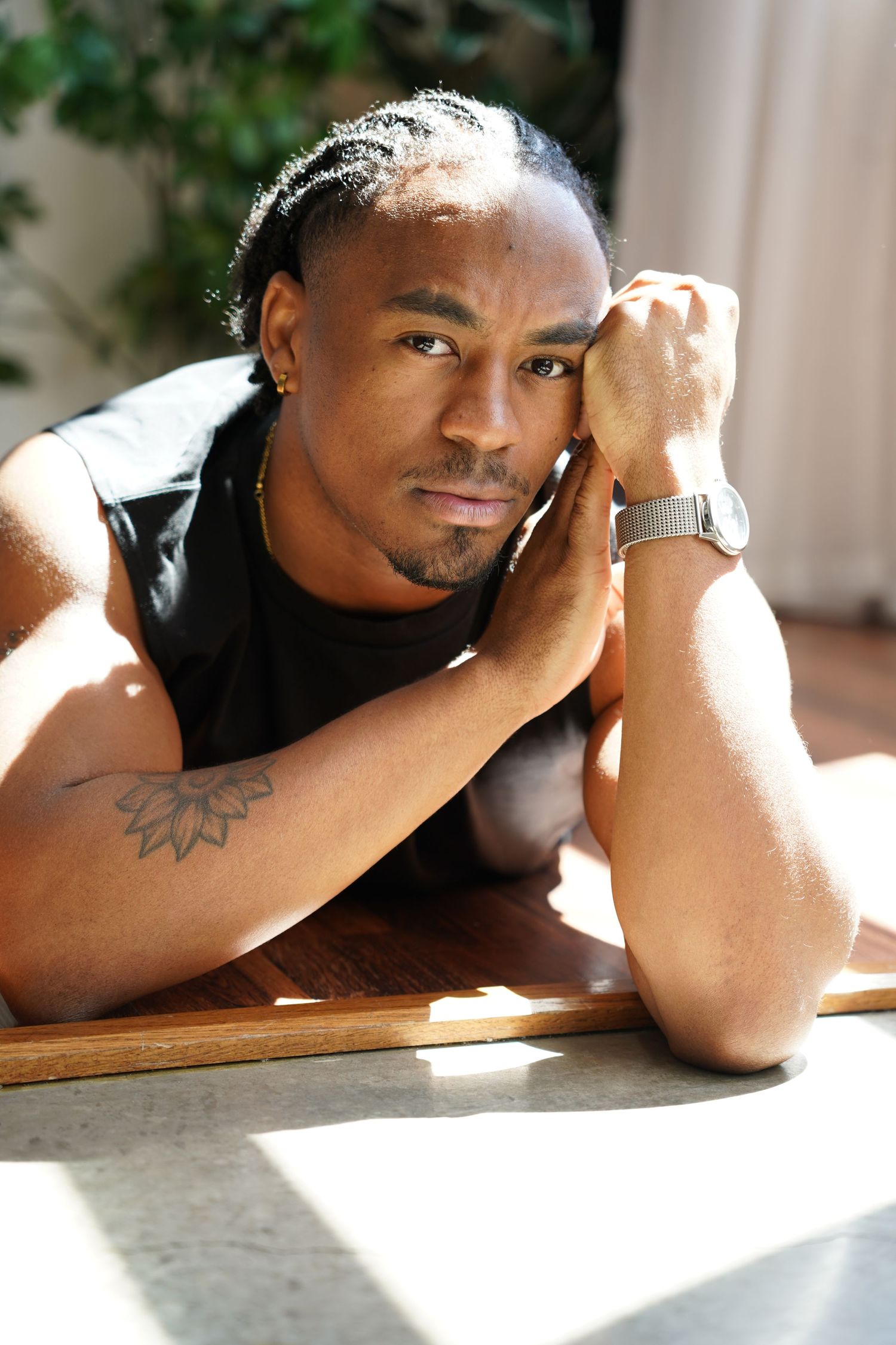 A muscular Black man with braids and a tattoo rests his chin on his hand at a wooden table, wearing a watch.