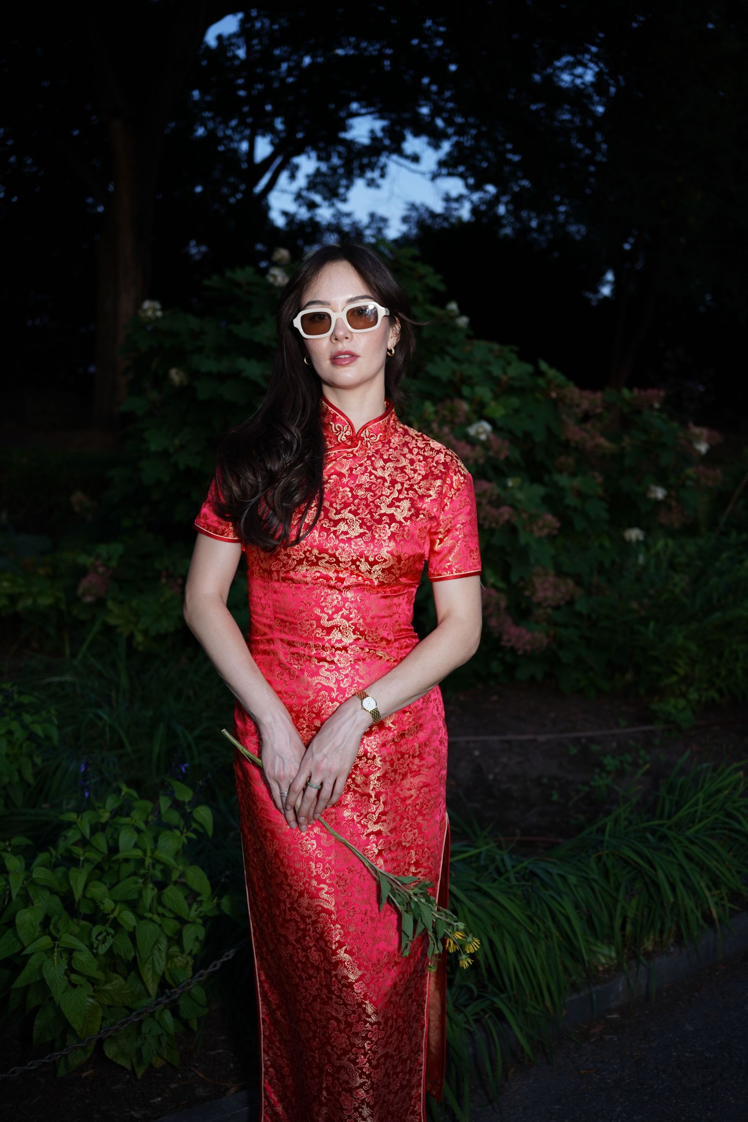 Woman in red floral cheongsam dress wearing white sunglasses, holding flowers in a lush garden setting.