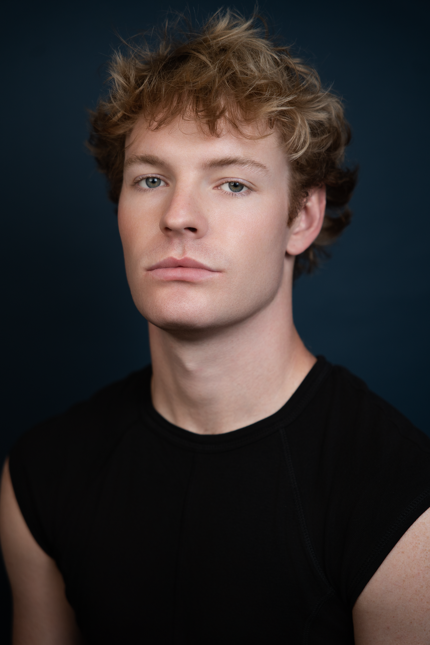 Young man with curly blonde hair and blue eyes wearing a black sleeveless shirt against a dark blue background.