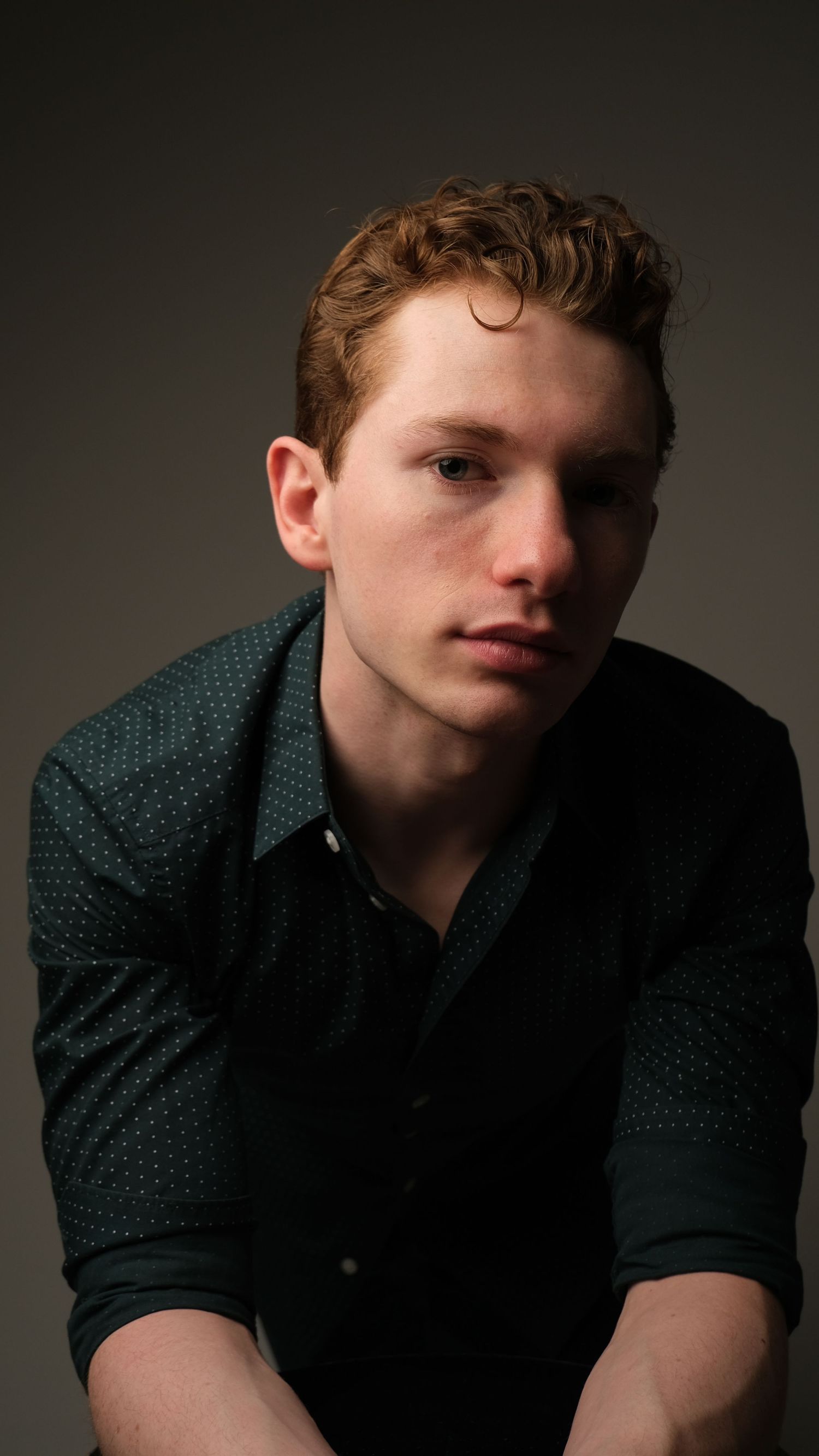 Portrait of young person in dark green patterned shirt against moody backdrop with dramatic lighting.