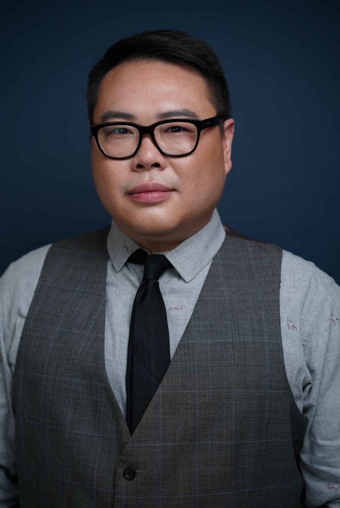 Professional headshot portrait of someone wearing glasses, a black tie and grey vest against a dark background.