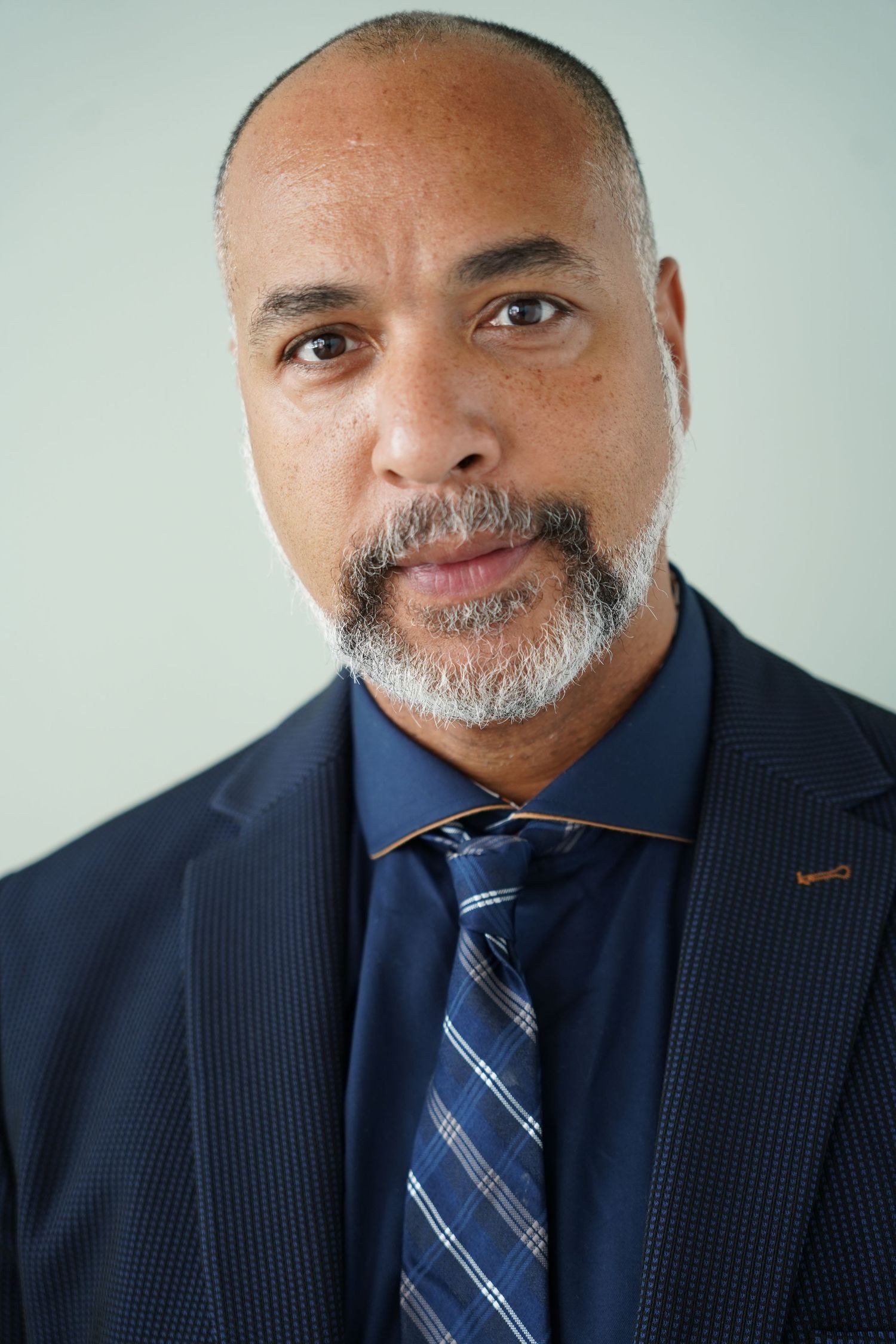 Professional headshot of a middle-aged man in a navy suit and plaid tie with a short gray beard.