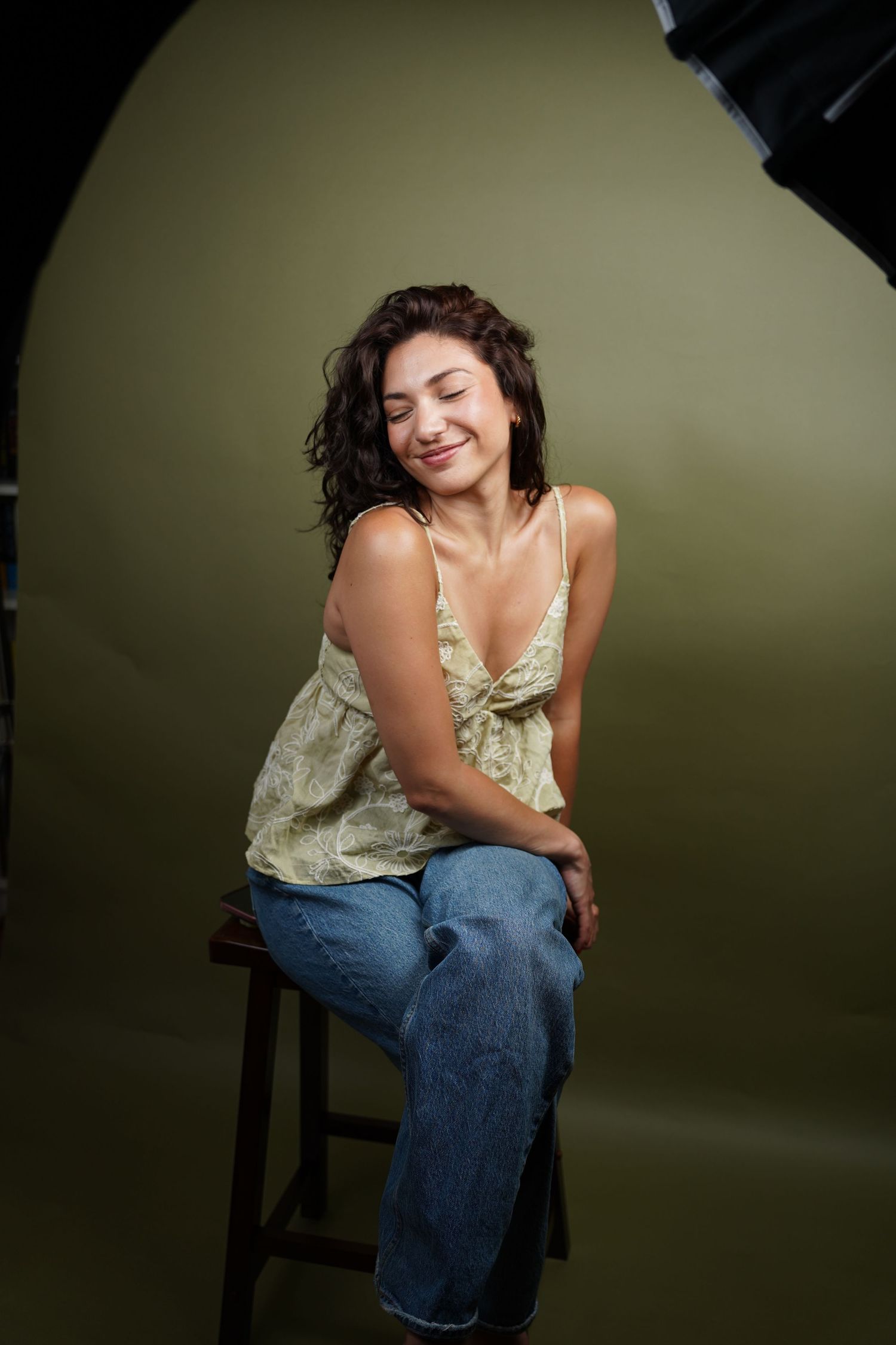 Smiling young woman with curly hair sitting on a stool in a photography studio, wearing a floral top and jeans.