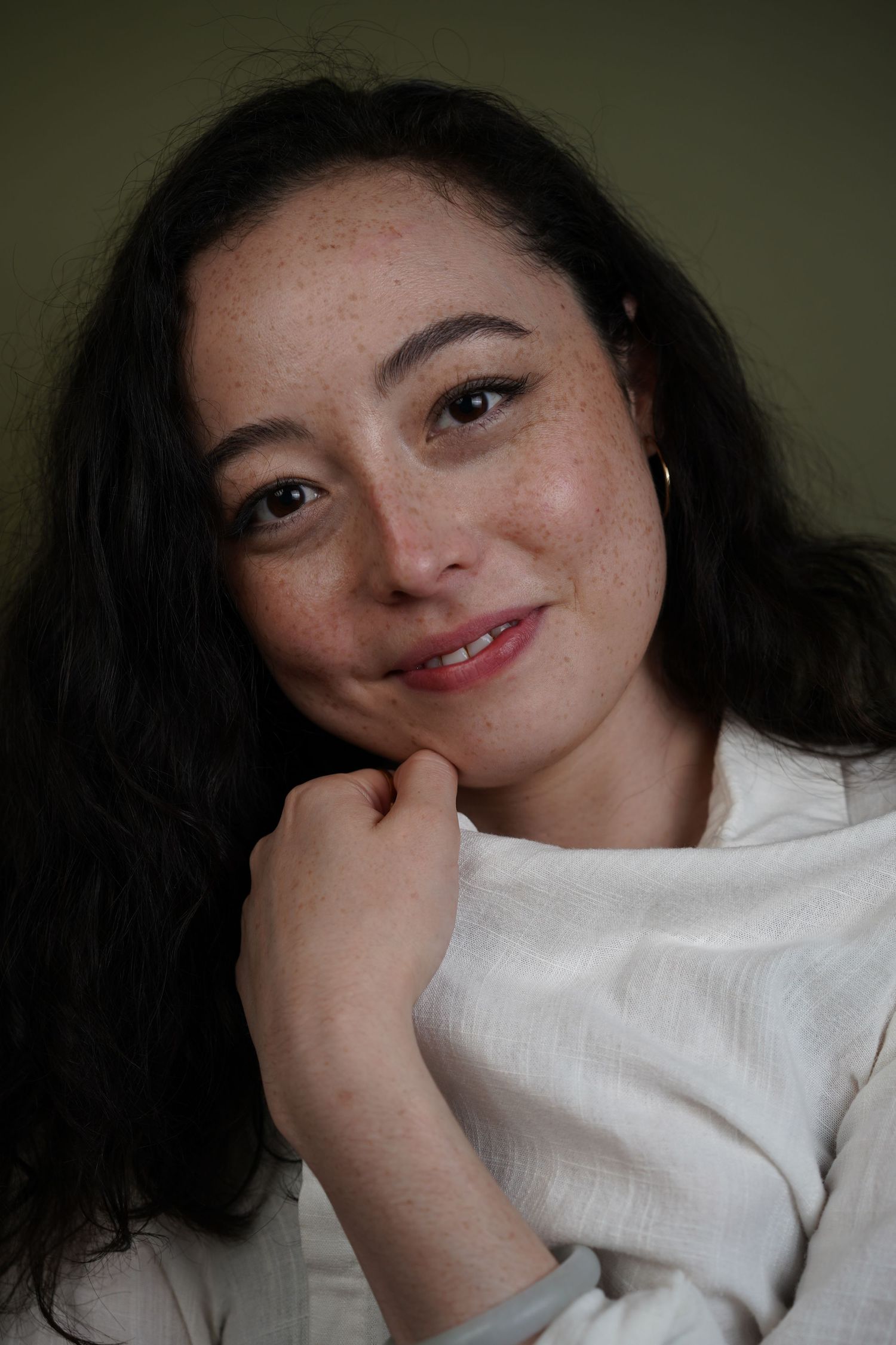 Portrait of someone with dark wavy hair wearing a white shirt, smiling warmly against a dark background.