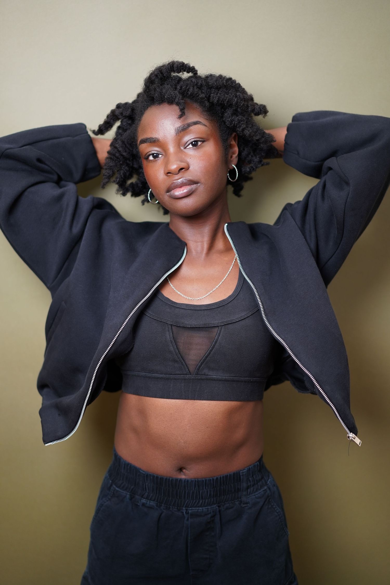 Young Black woman in black crop top and zip-up jacket poses confidently with hands behind head against olive background.
