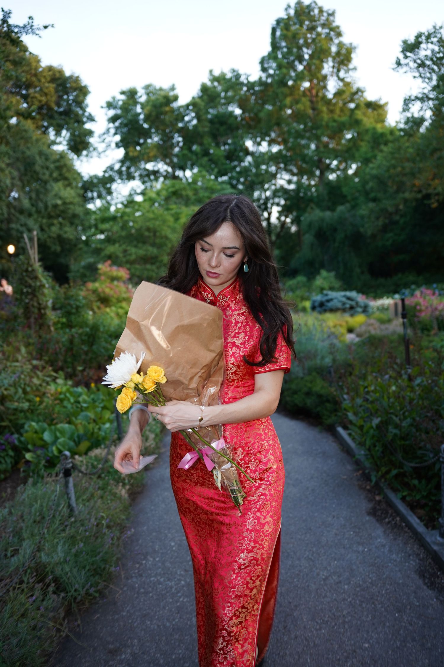 Woman in red cheongsam dress holding bouquet of yellow and white flowers on garden path.