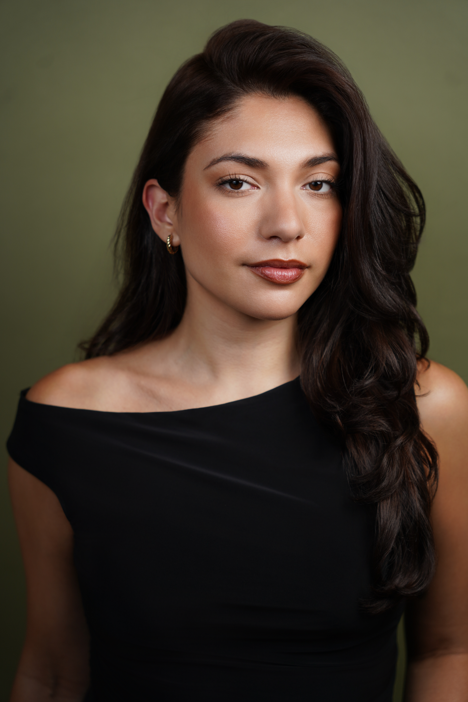 Young woman with long dark hair wearing a black off-shoulder top poses for a professional portrait.