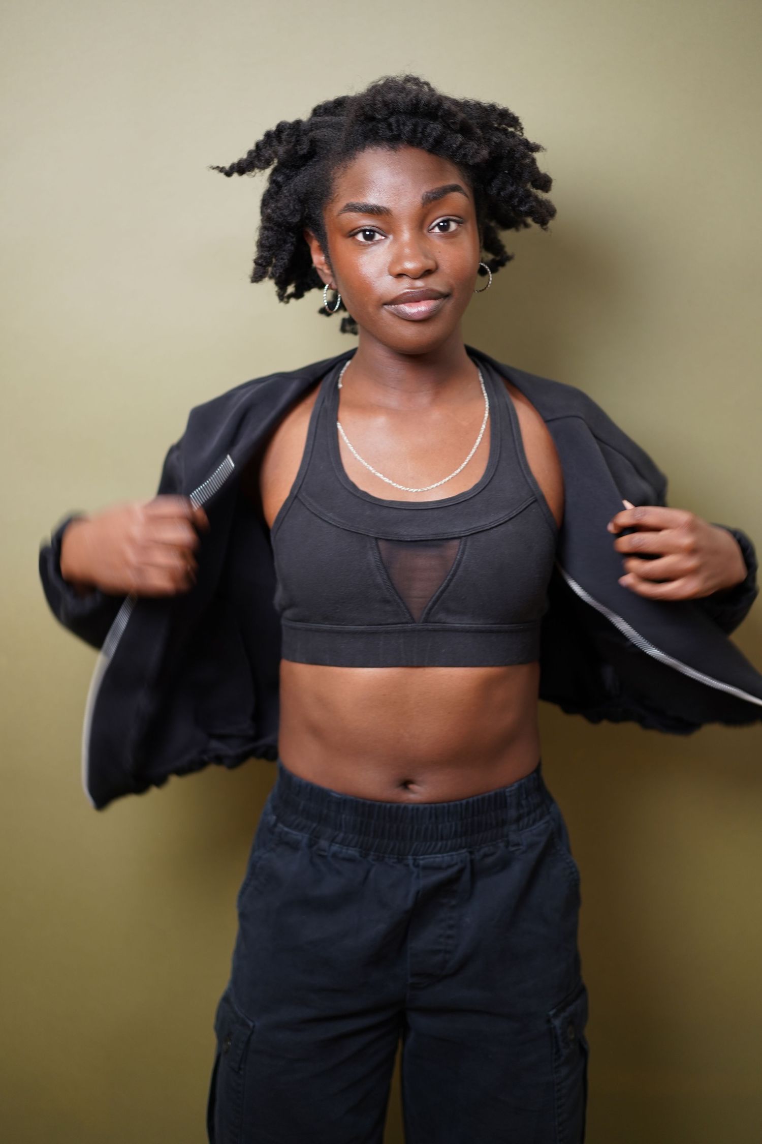 Young Black woman in black sports bra and cargo pants, opening jacket, posing confidently against olive green background.