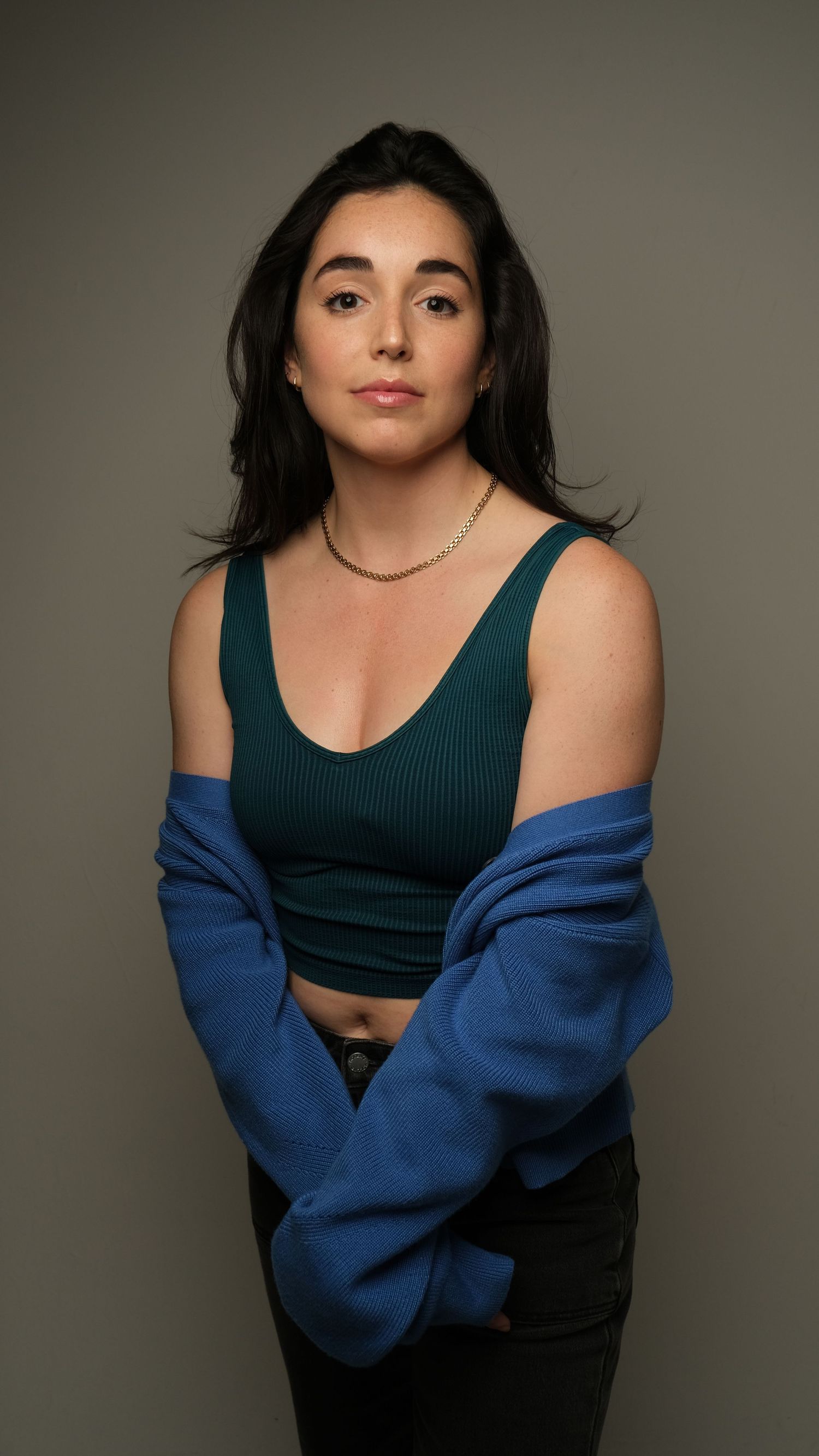 Portrait in a dark green crop top and blue sweater against neutral studio backdrop, showcasing casual style.