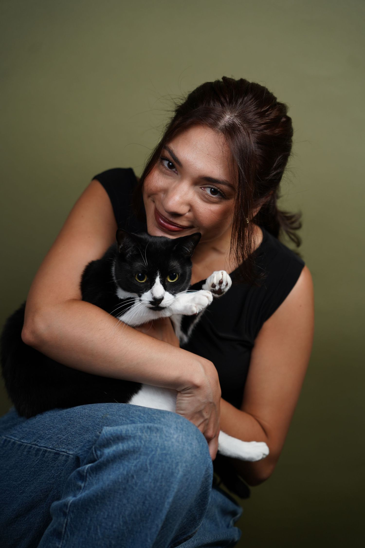 A smiling woman in a black top and jeans holds a black and white cat close to her face against a green wall.