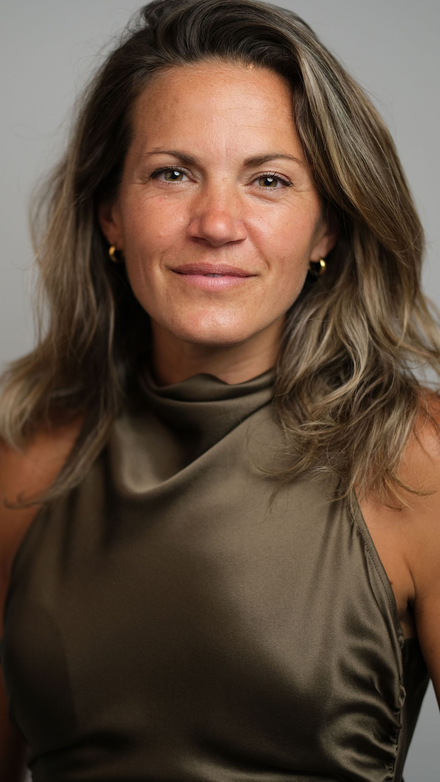 Professional headshot portrait of a person with wavy brown hair wearing an olive sleeveless top against a neutral background.