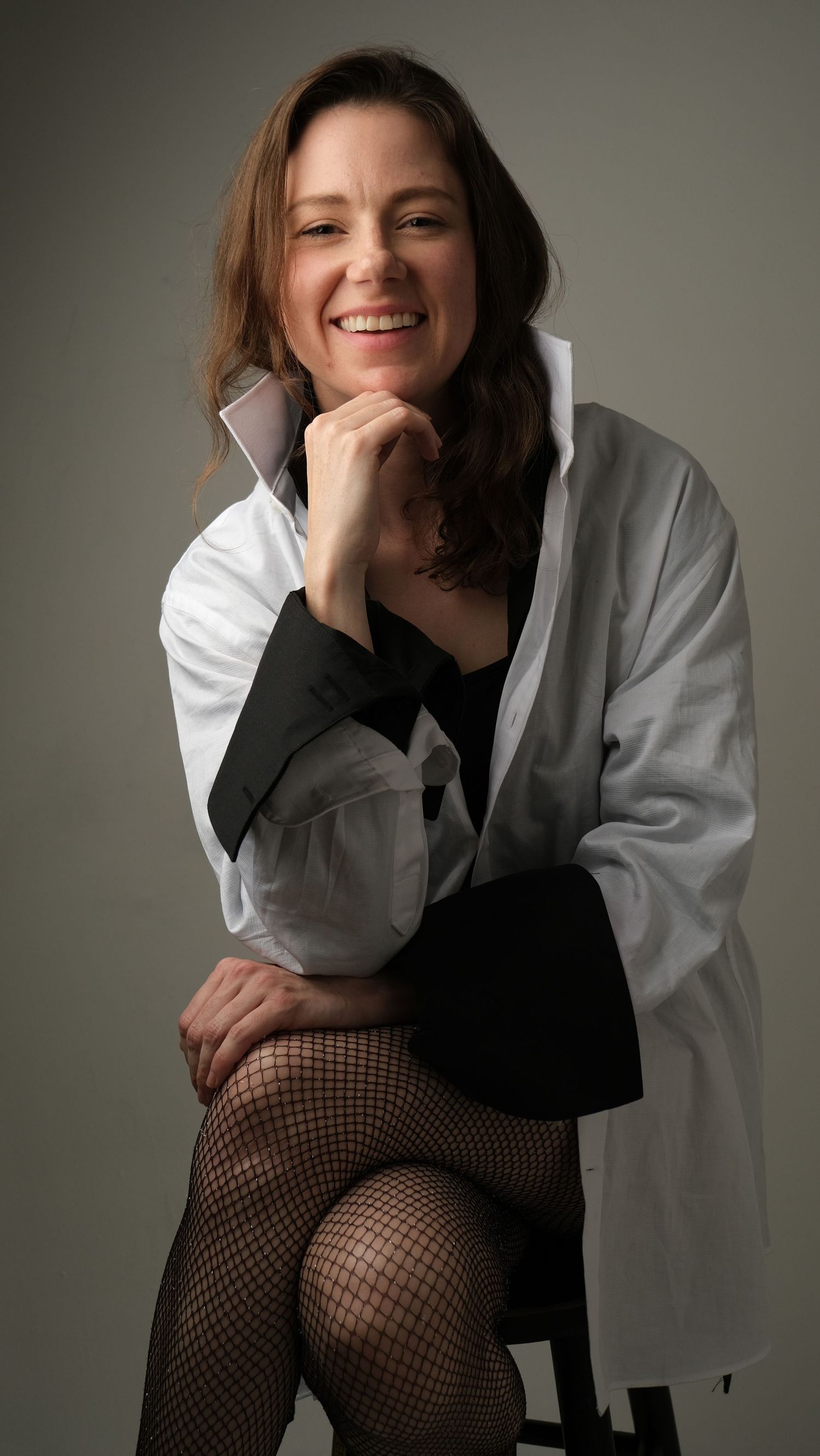 Professional portrait of a woman in white jacket and black dress with fishnet stockings seated against gray backdrop.