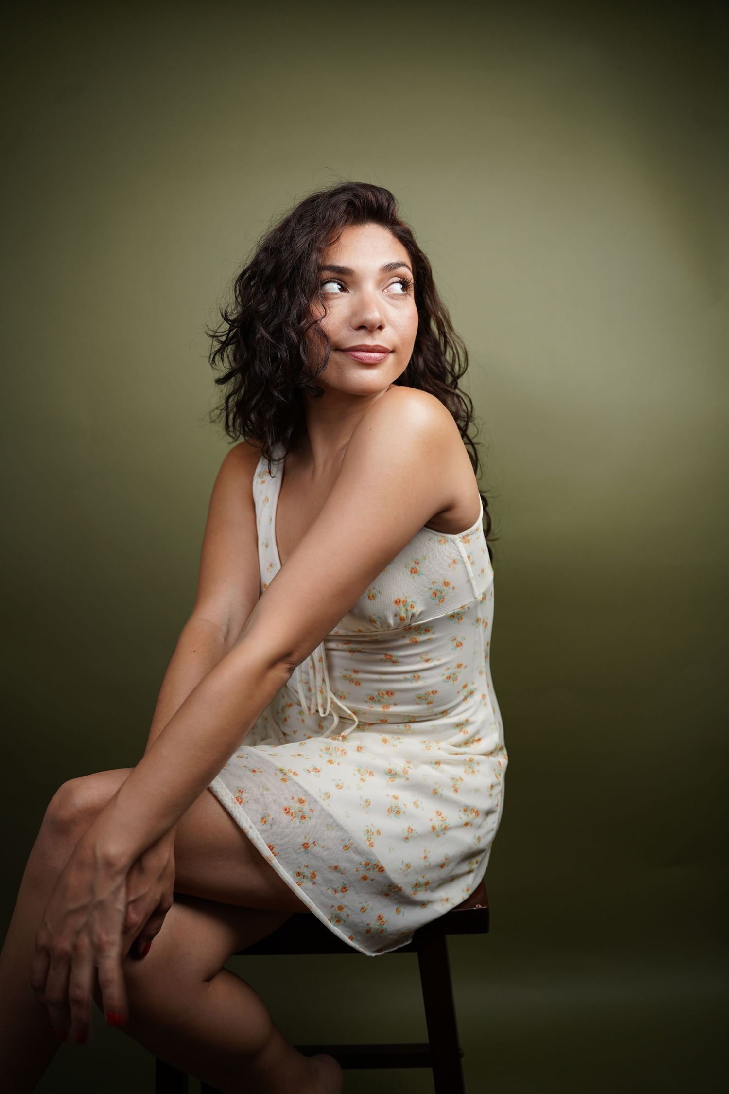 Young woman in floral sundress sits on stool, gazing upward thoughtfully against olive green background.