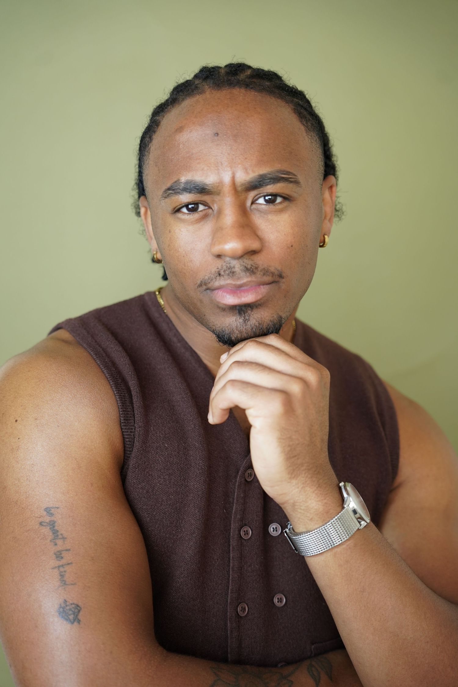 Young Black man with braids, goatee, tattoos, and a watch poses thoughtfully with hand on chin against green background.