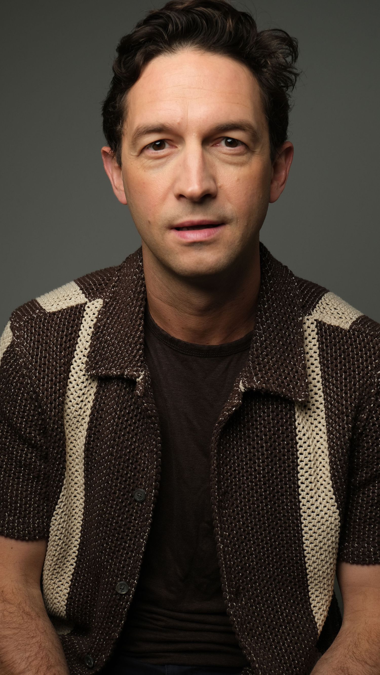 Professional studio portrait of someone wearing a dark textured vest with beige stripes against a dark background.