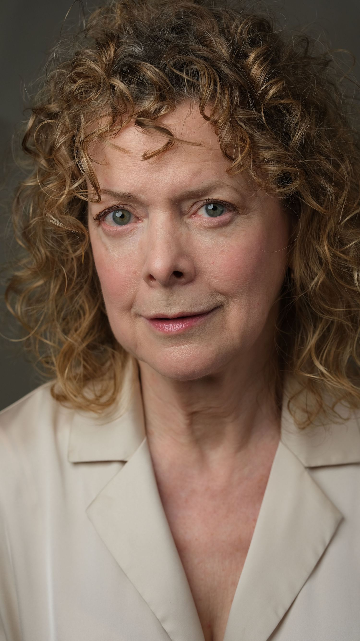 Professional headshot portrait featuring curly blonde hair and a cream colored blazer against a dark background.
