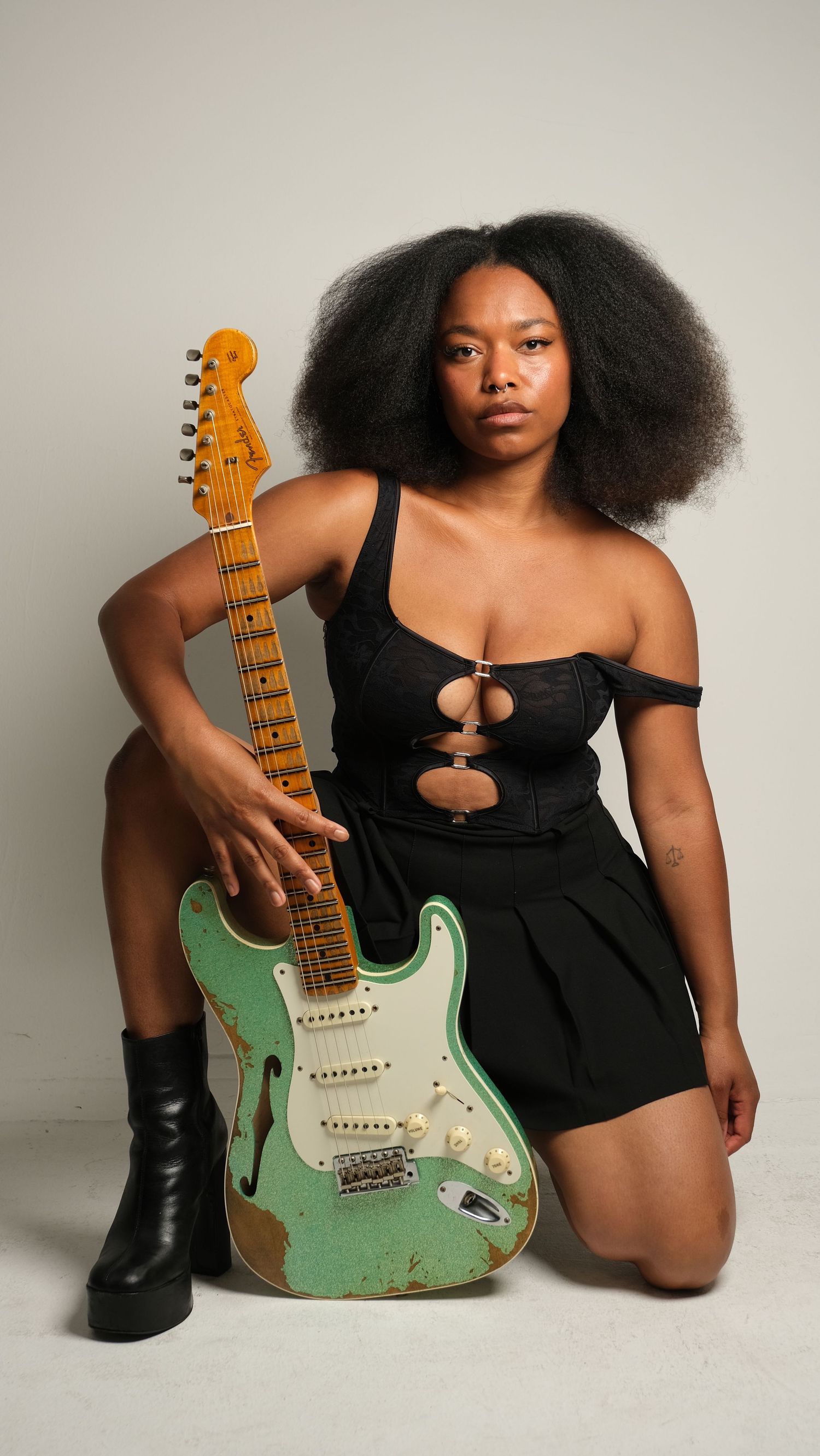 Musician poses with vintage mint green Fender Stratocaster electric guitar wearing black outfit and boots against neutral background.