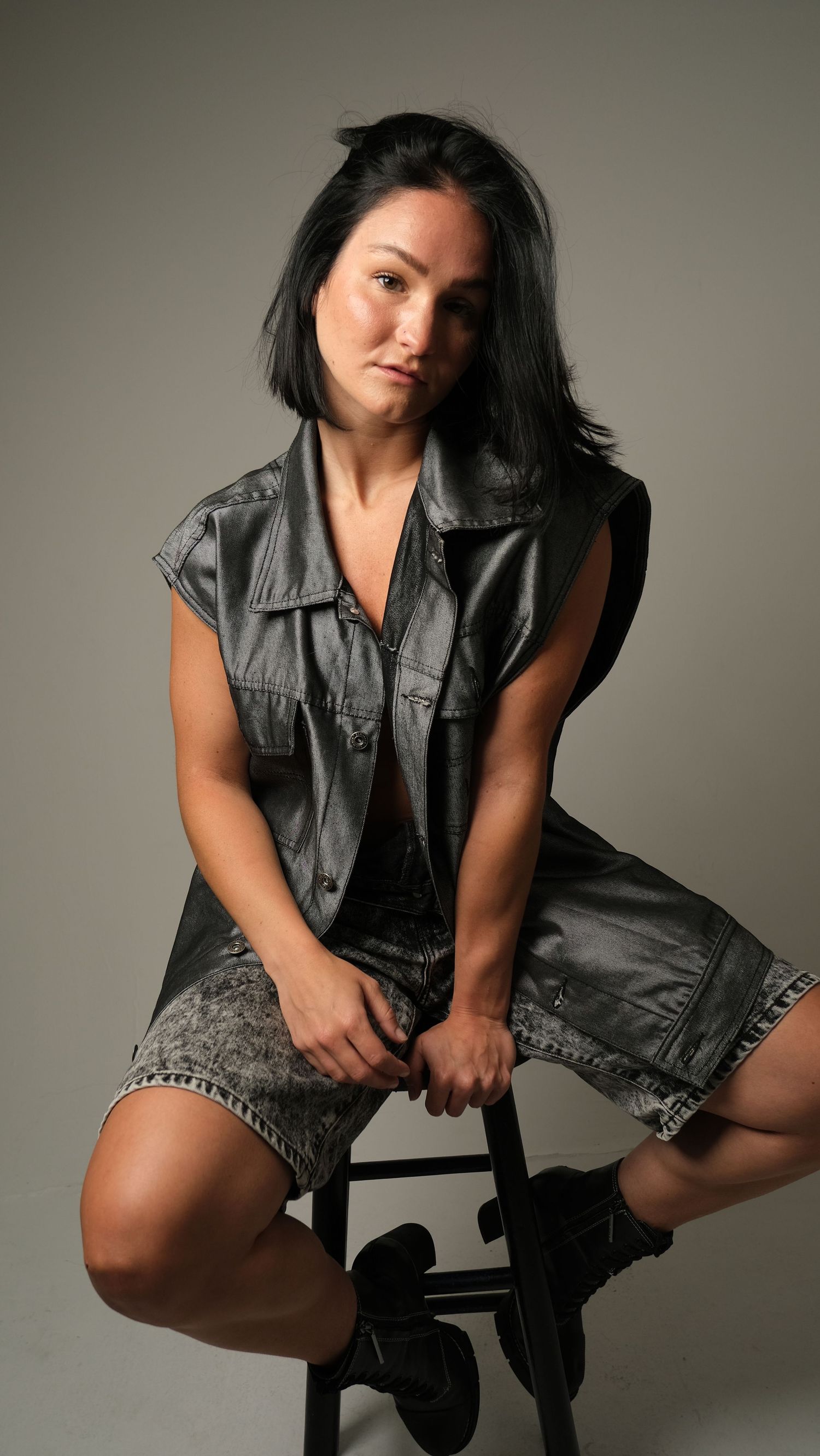 Model in grunge style outfit featuring dark denim vest and shorts sits on stool in studio with neutral backdrop.